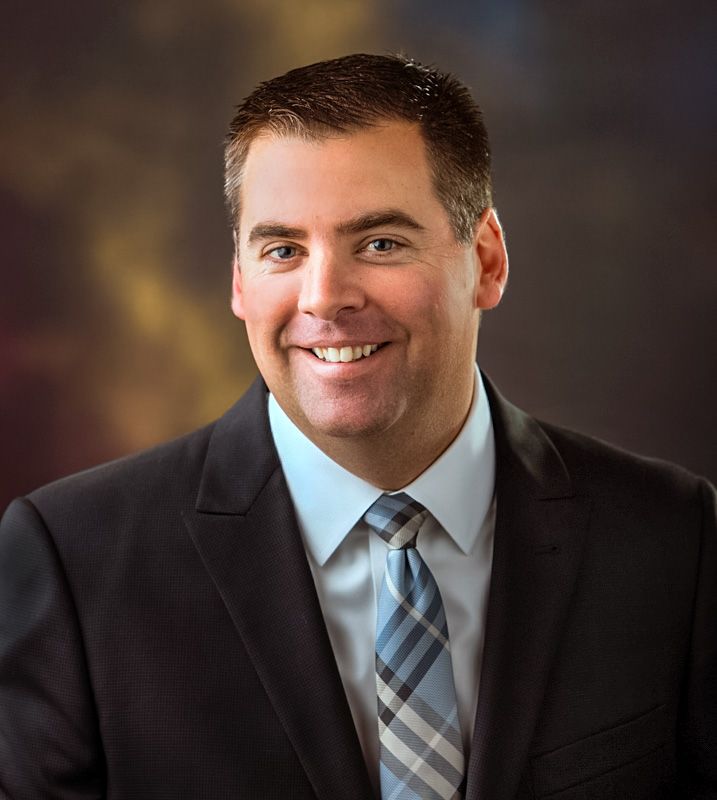 Professional headshot of a smiling man in a dark suit and plaid tie against a warm bokeh background.