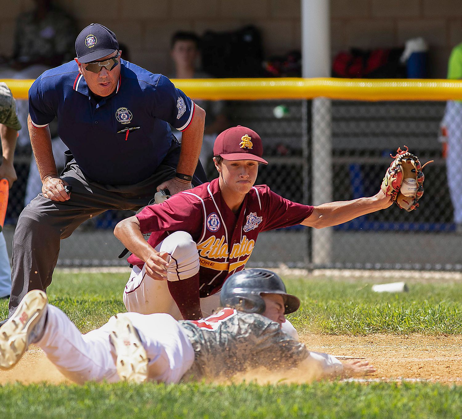 Baseball player sliding into base as fielder catches ball with umpire watching closely during a youth league game.
