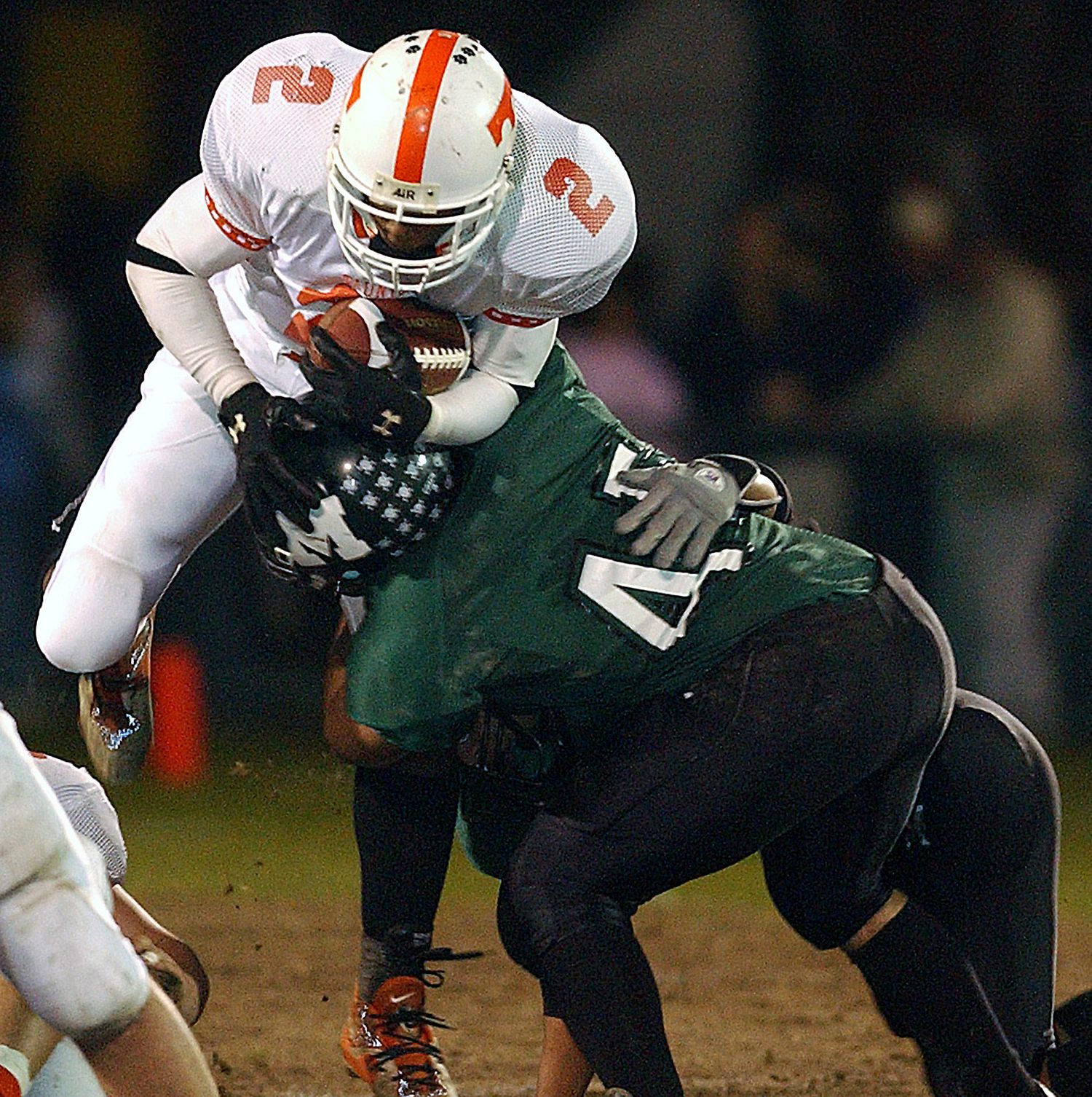 Football player in white jersey number 2 tackled by green jersey player 47 during intense night game action.