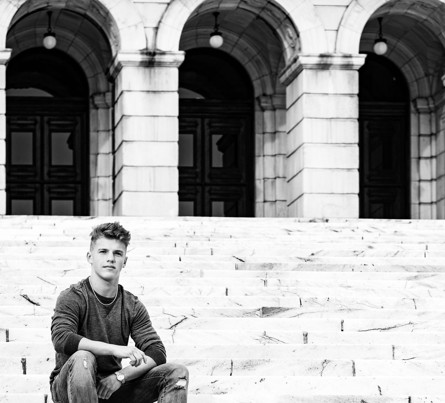 Young man sitting on stone steps in front of a grand arched building facade, black and white photo.