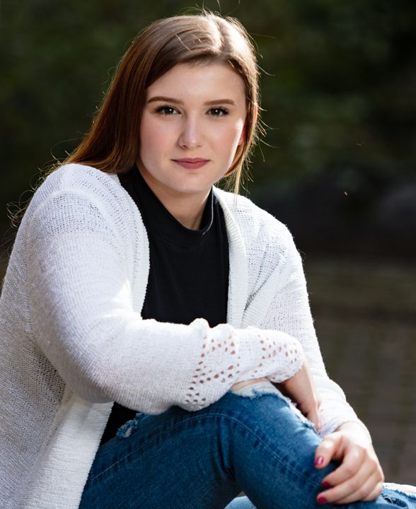 Young woman with auburn hair in white cardigan and jeans poses outdoors for a senior portrait photo.