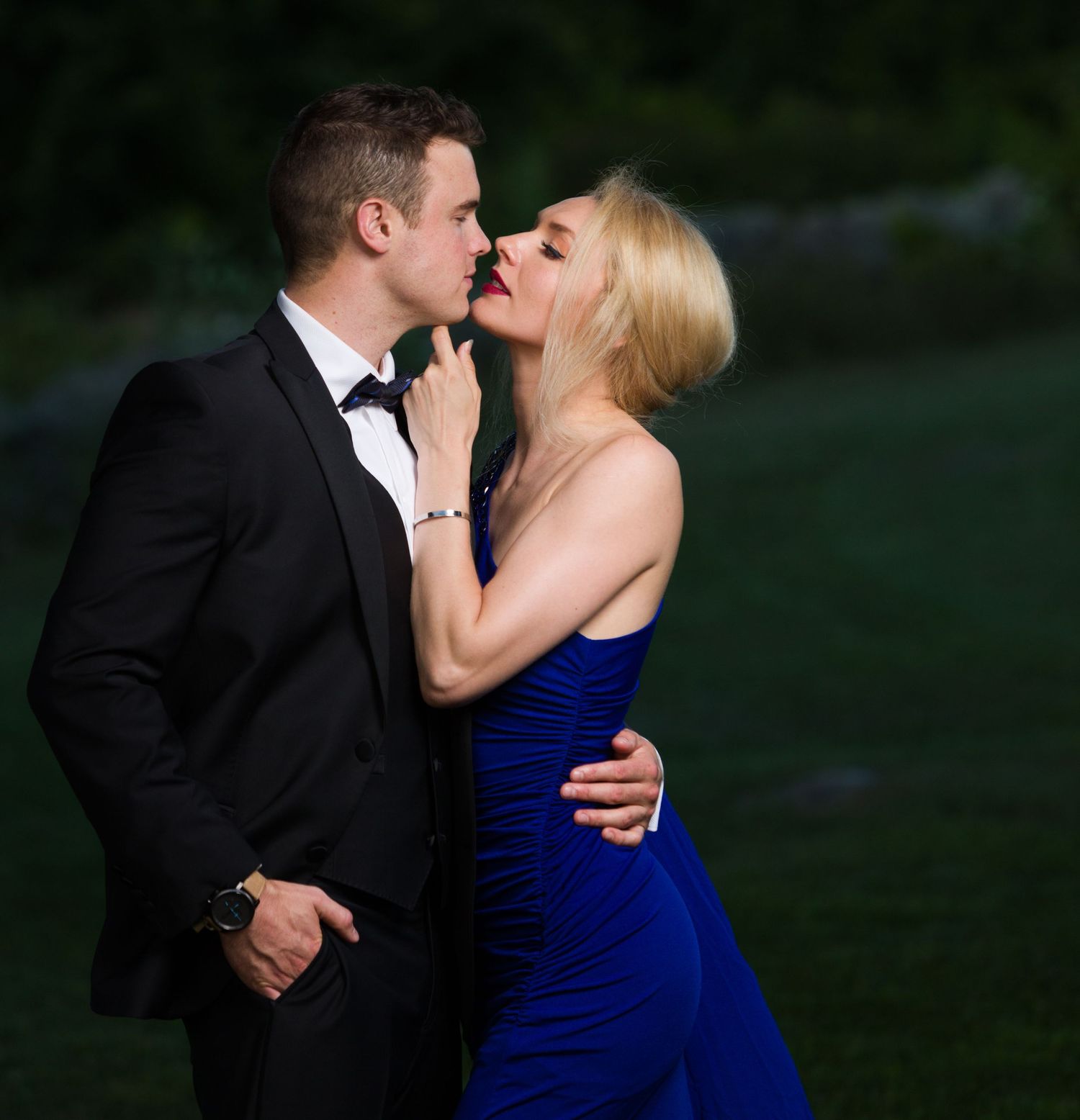 Couple in formal wear sharing a romantic kiss outdoors, man in black tuxedo and woman in royal blue gown.