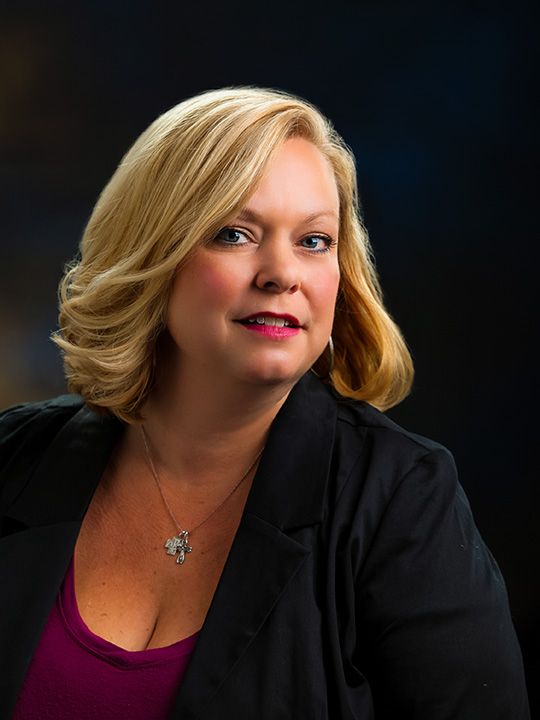A professional headshot of a blonde woman in a black blazer against dark background.
