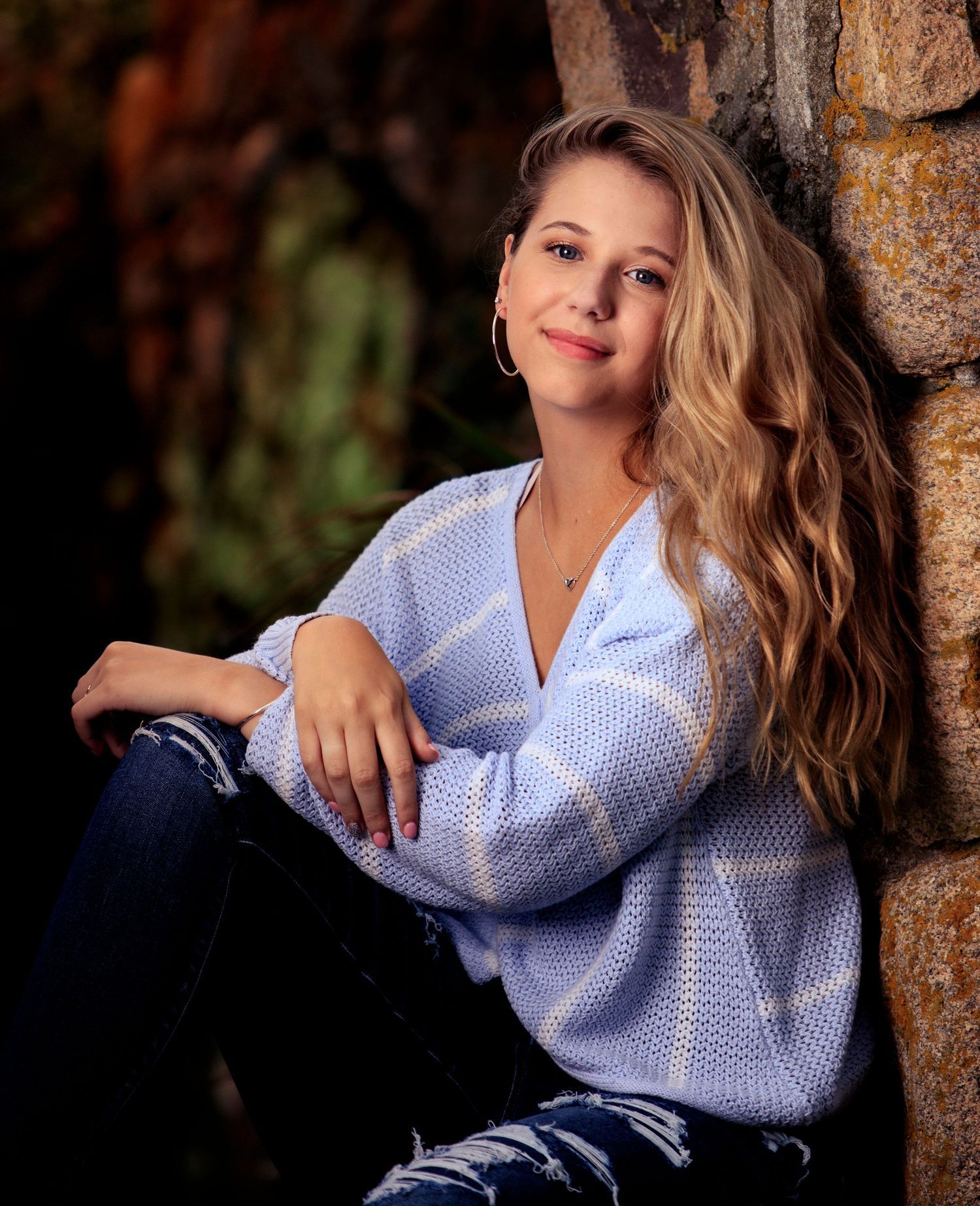 Young woman with wavy blonde hair in a light blue sweater and jeans, leaning against a stone wall outdoors.