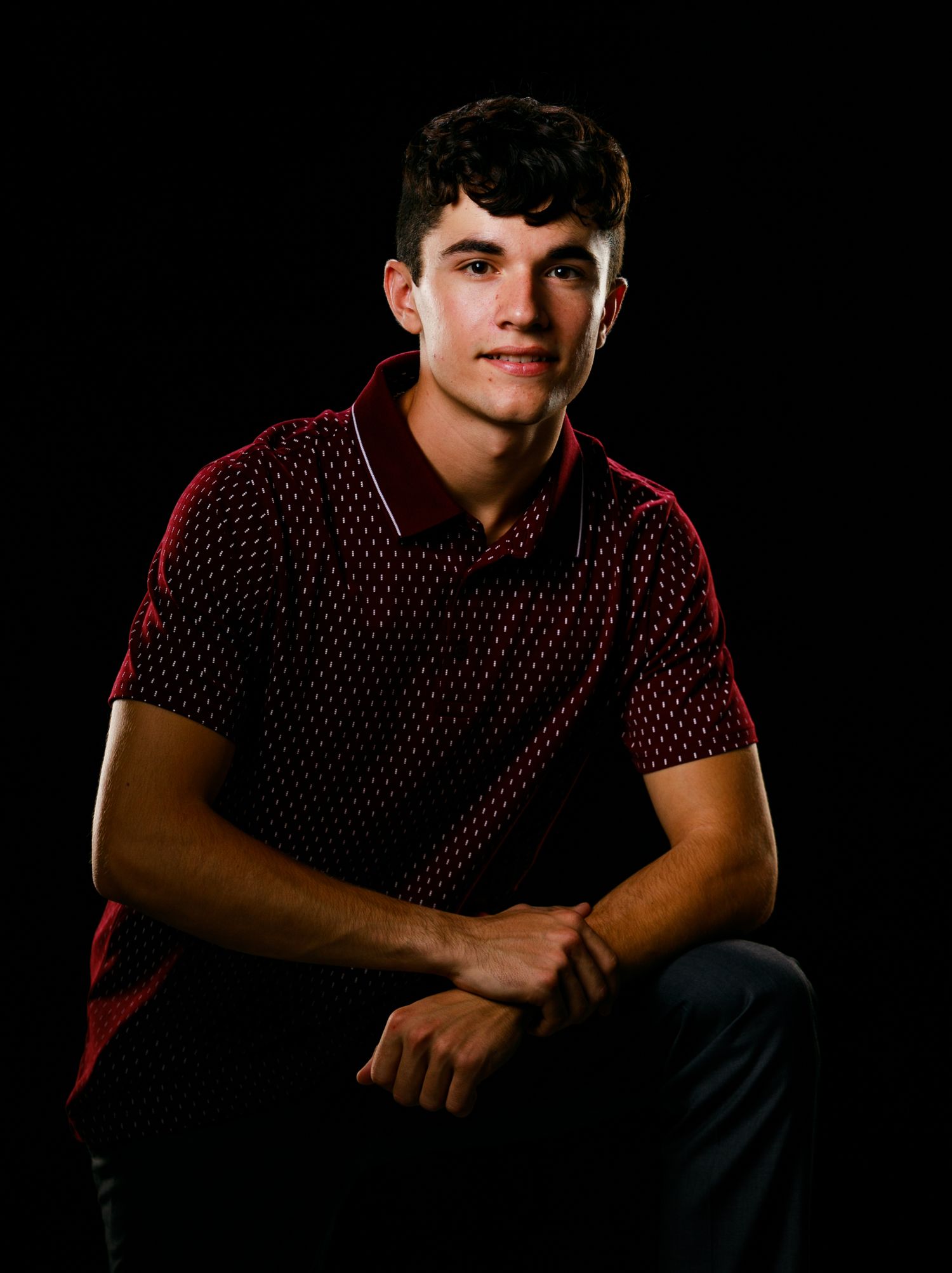 Young man in dark red patterned polo shirt seated against black studio background portrait.