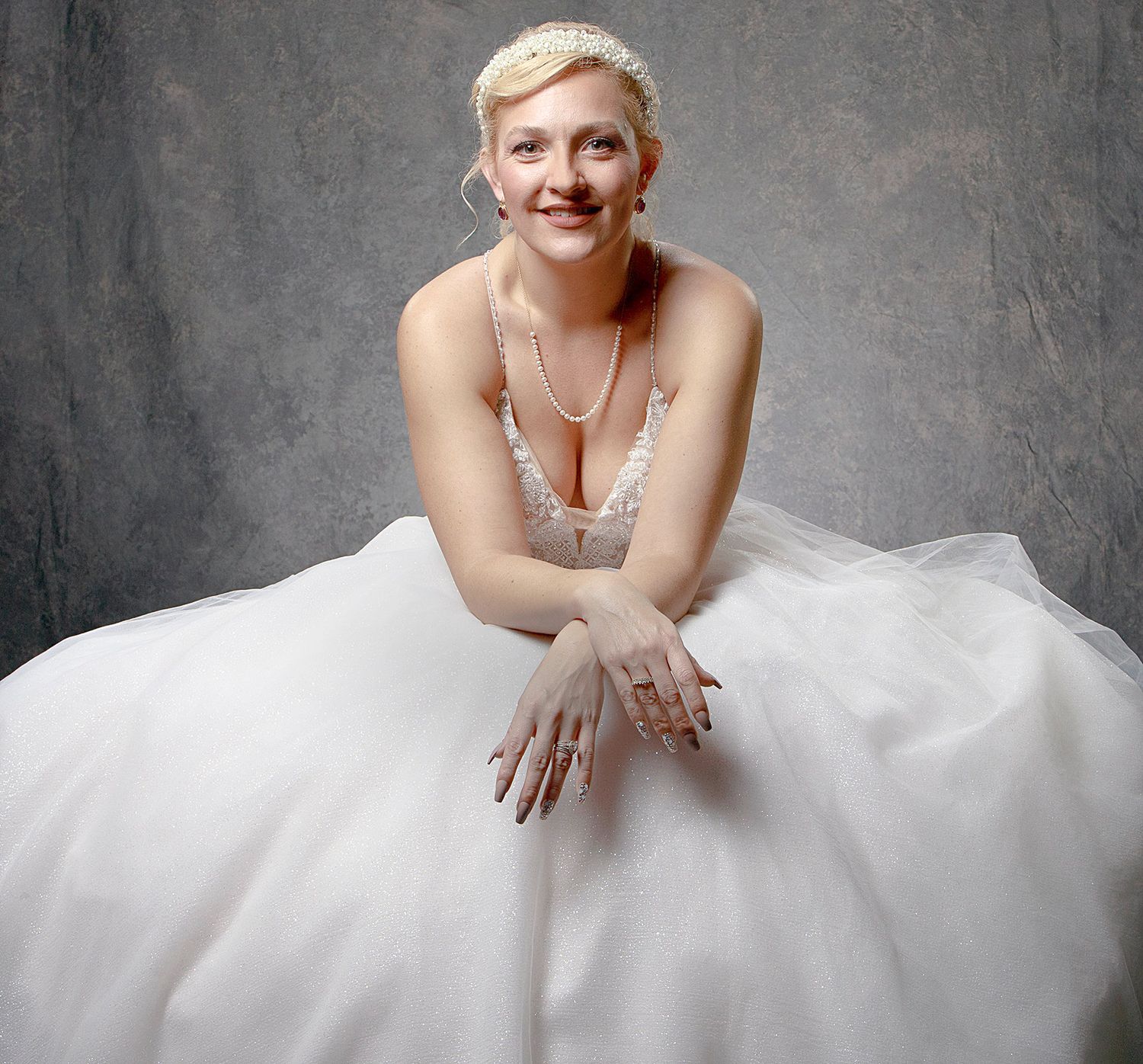 Smiling blonde bride in a white ballgown with pearl necklace poses elegantly against a gray studio background.
