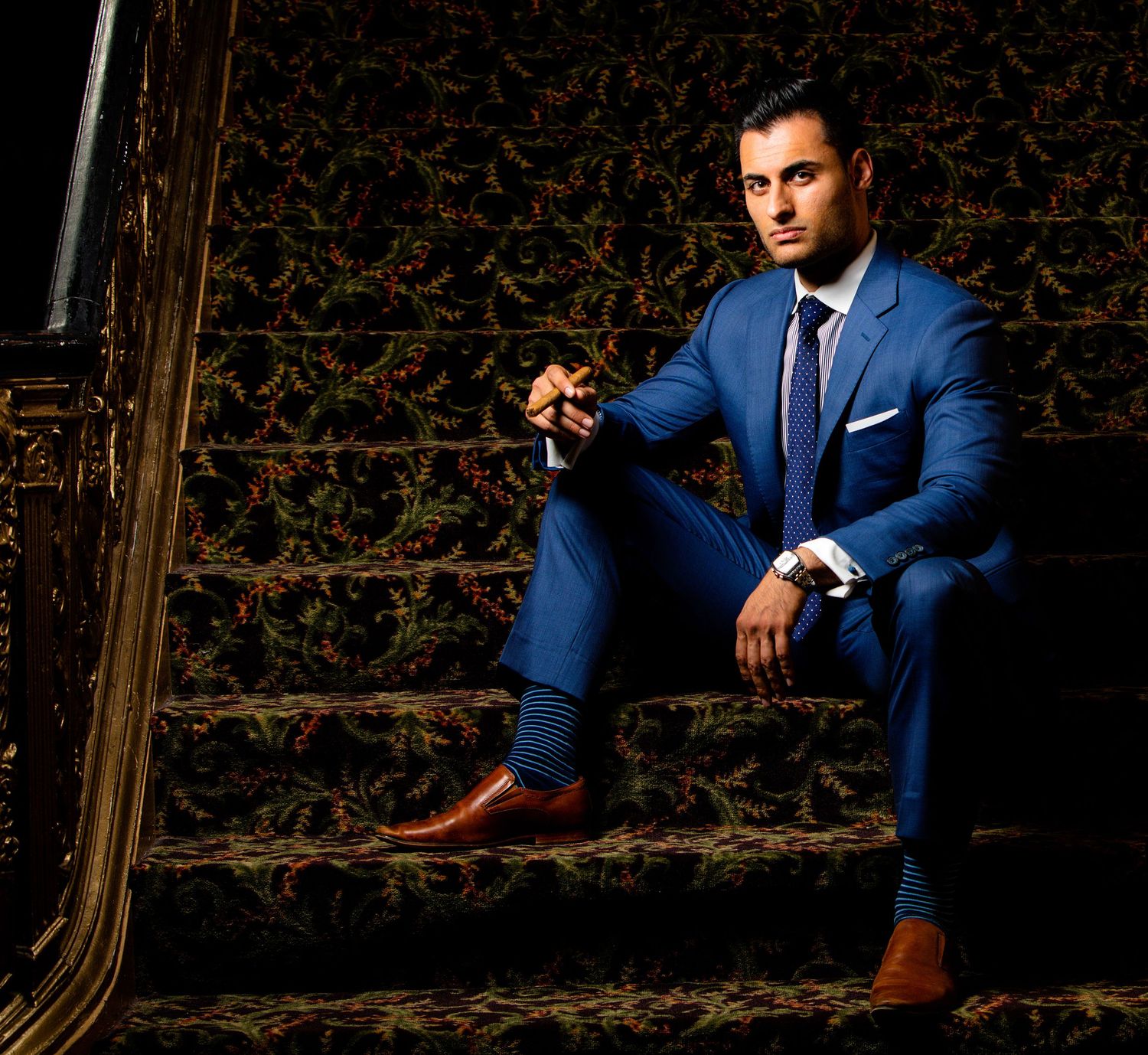 Man in blue suit and brown shoes sitting on ornate carpeted stairs in a professional portrait photo.