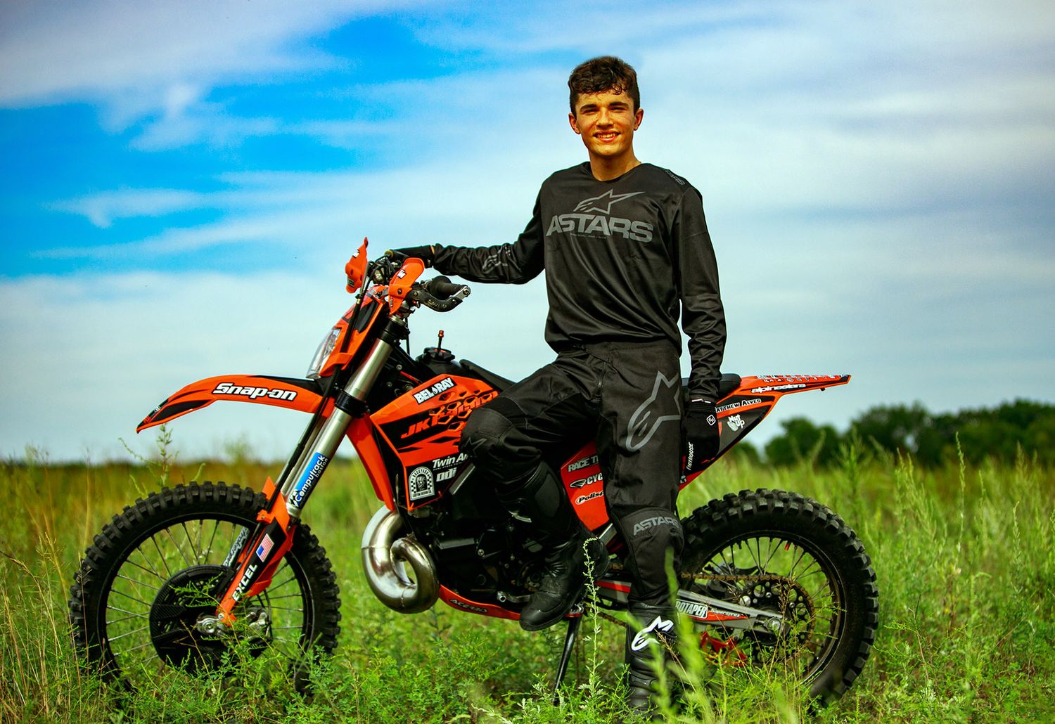 Young man in motocross gear posing with orange dirt bike in green field under cloudy sky.