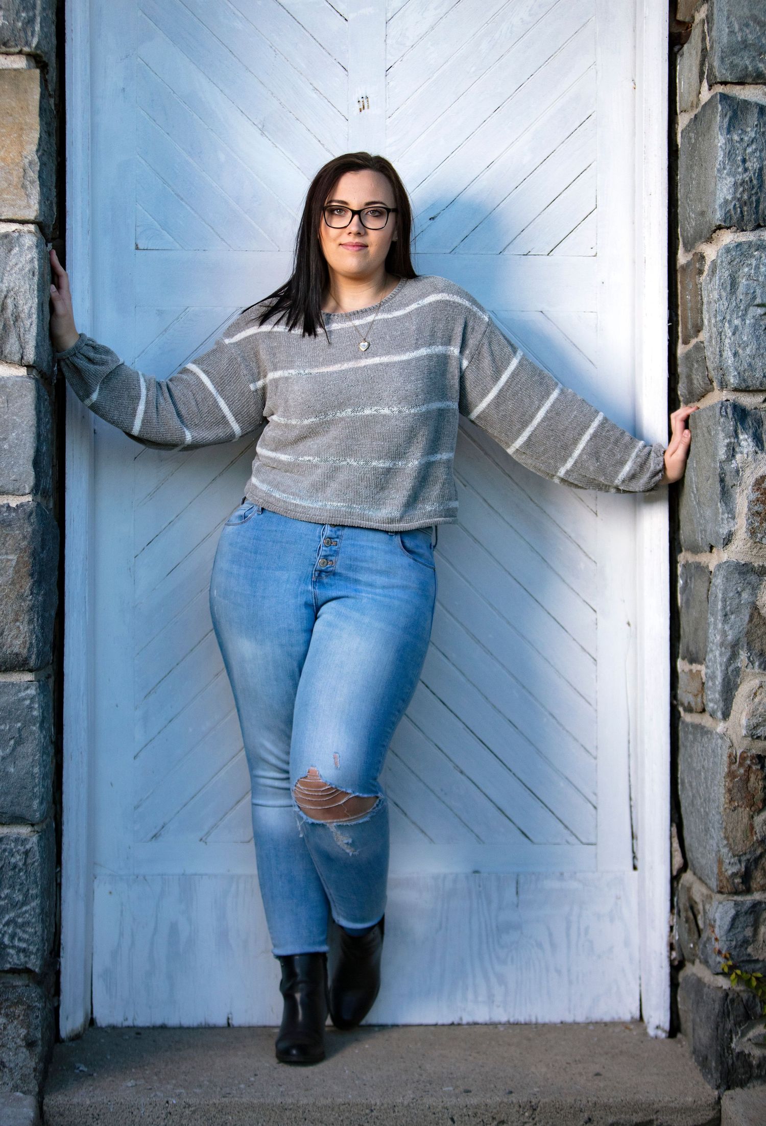 Young woman in striped sweater and ripped jeans poses in white wooden doorway of stone building.