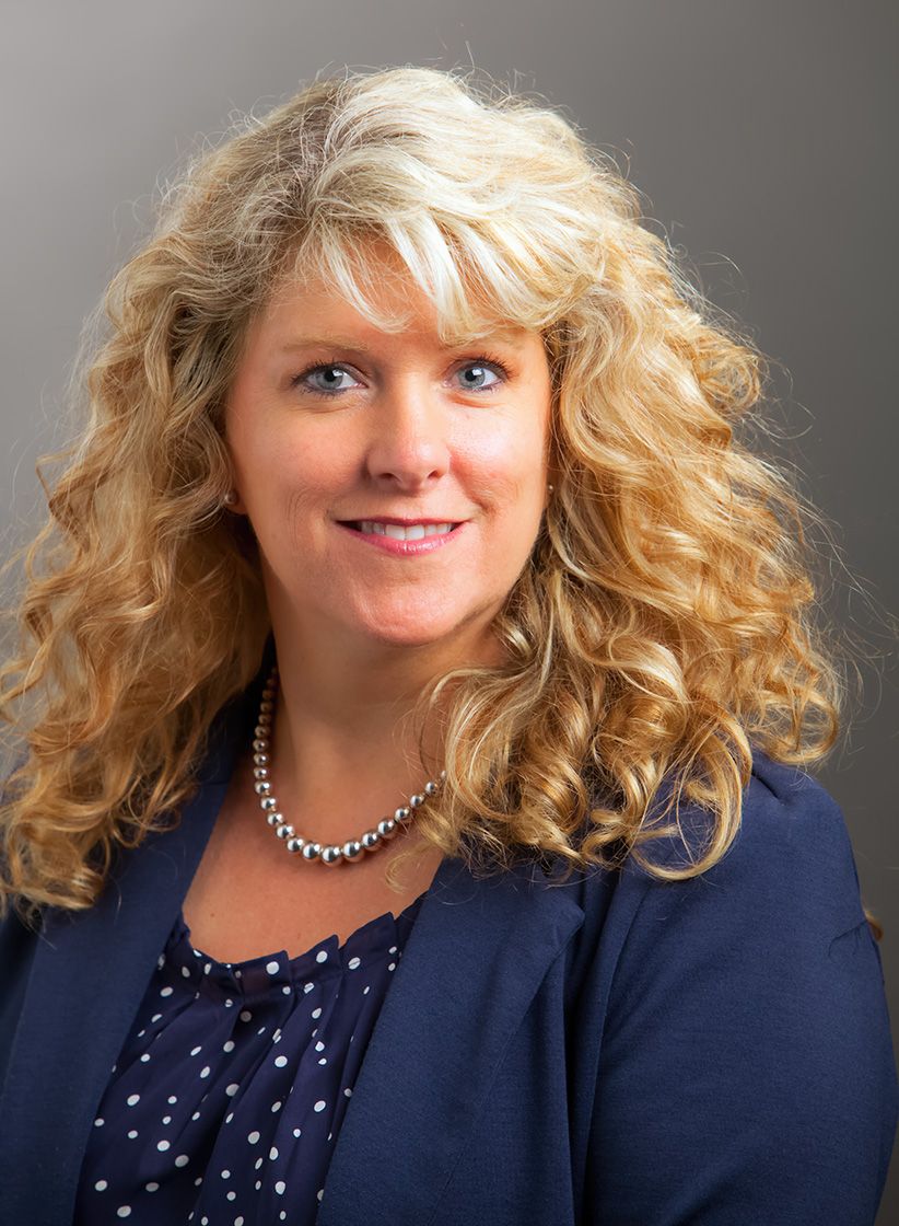 Professional headshot of a smiling blonde woman with curly hair wearing a navy blazer and pearl necklace.