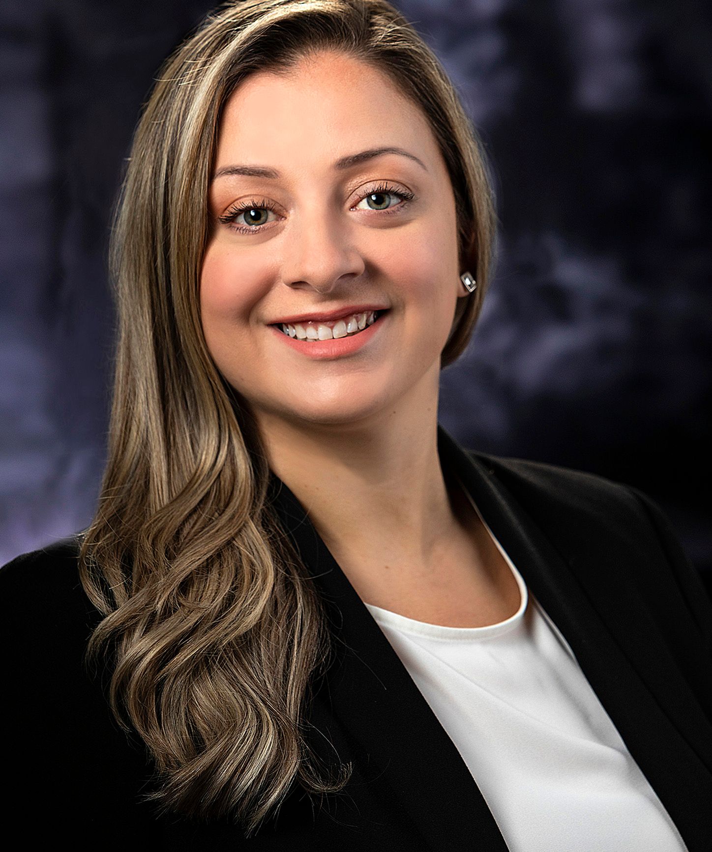Professional headshot of a smiling woman with highlighted hair wearing a black blazer against a dark background.