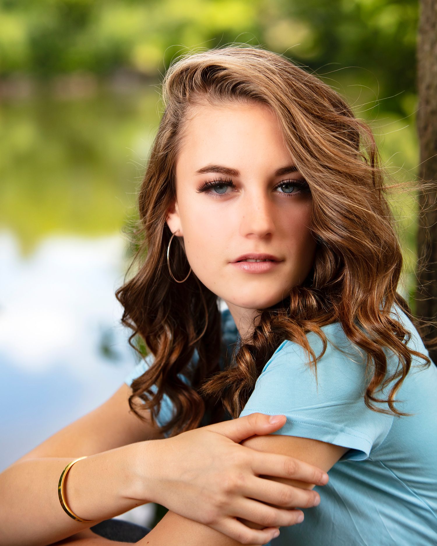 Young woman with curly brown hair wearing a light blue top and gold bracelet poses outdoors near water.
