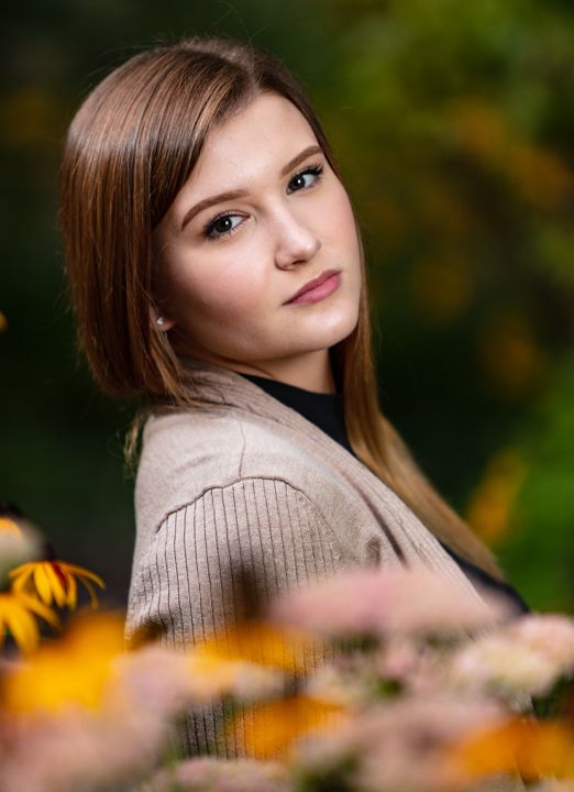 Young woman with straight brown hair poses outdoors among yellow flowers wearing a beige cardigan.