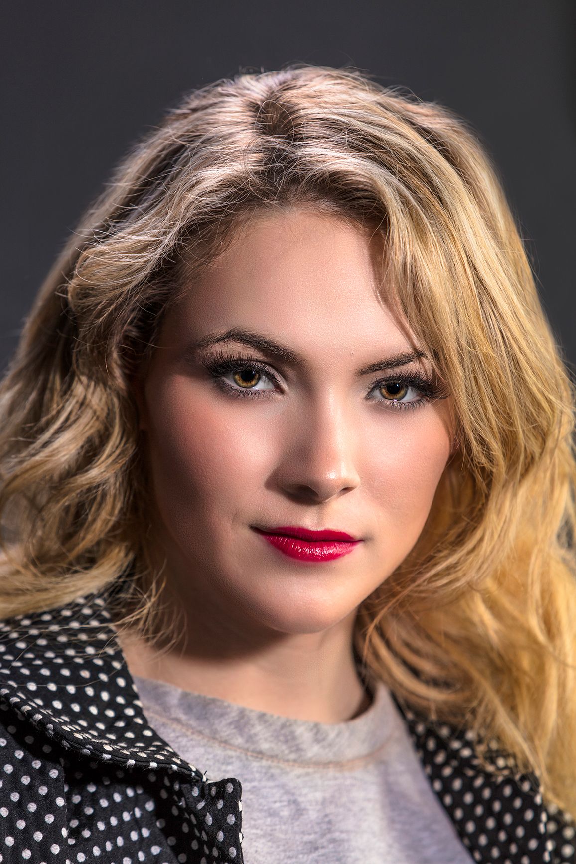 Blonde woman with red lips and smoky eye makeup wearing a polka dot jacket, posing against a dark gray background.