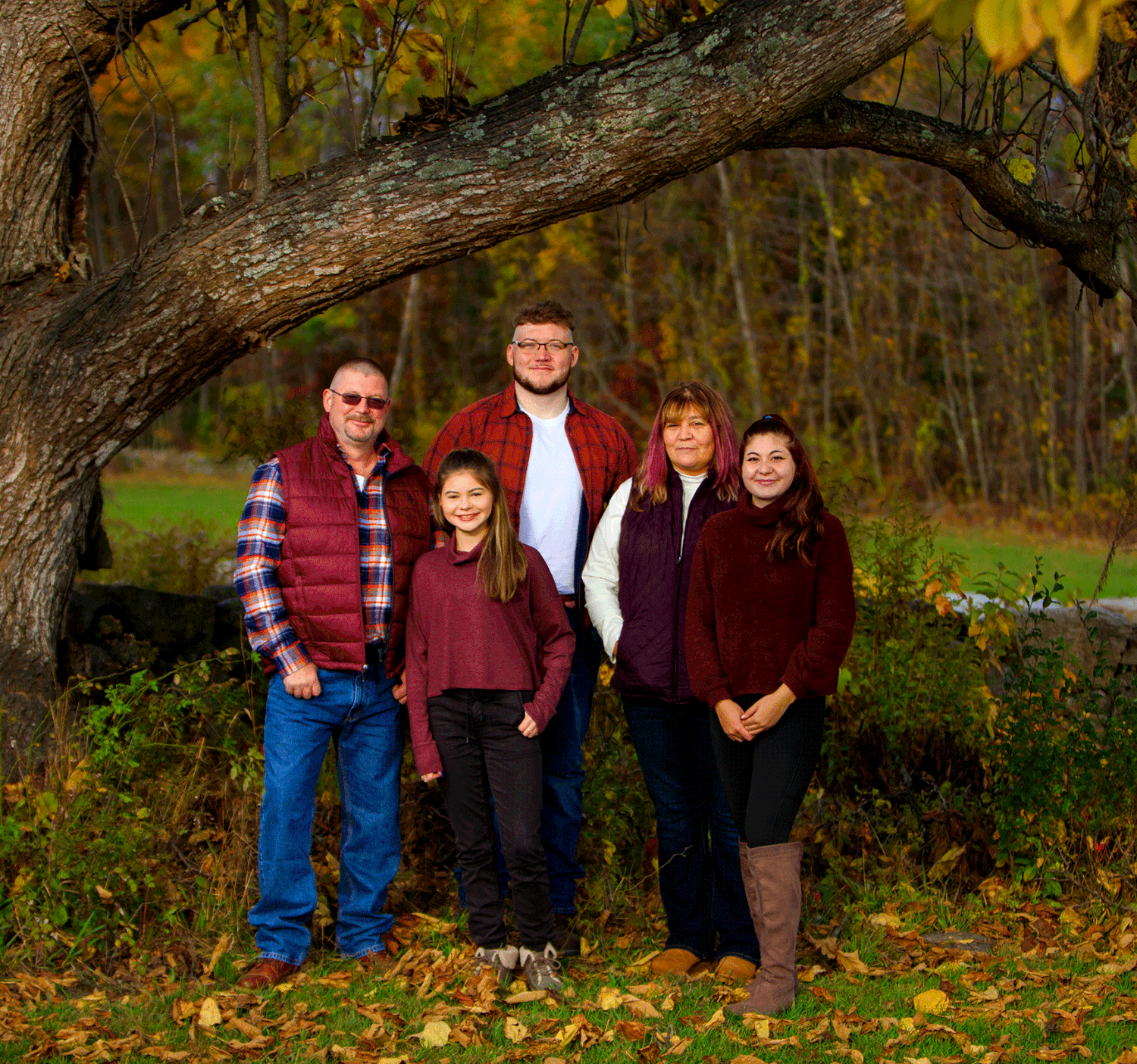 A family of five smiles together under a large tree during fall foliage season.
