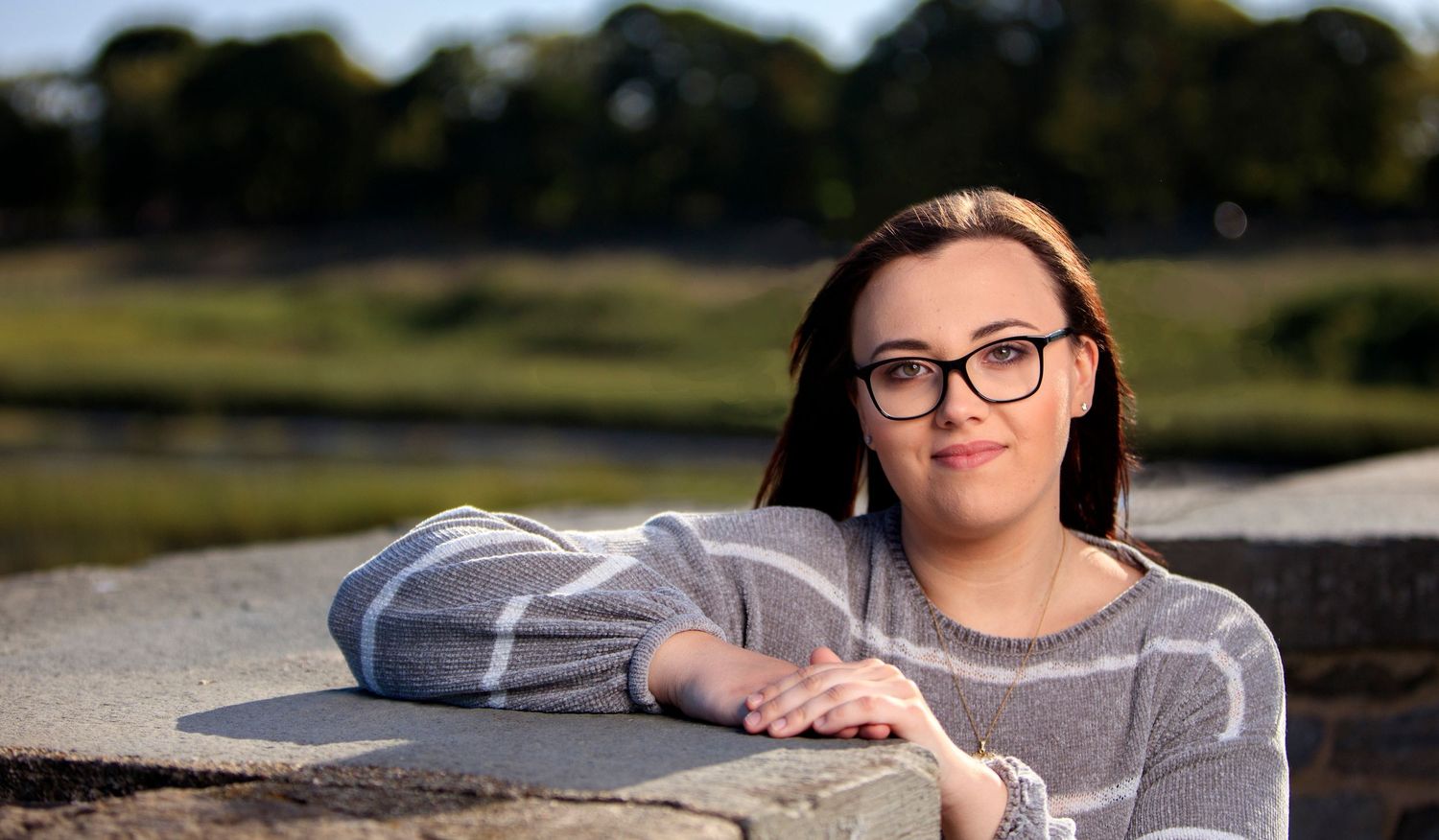 Young woman with glasses rests arms on stone wall outdoors near scenic waterway.