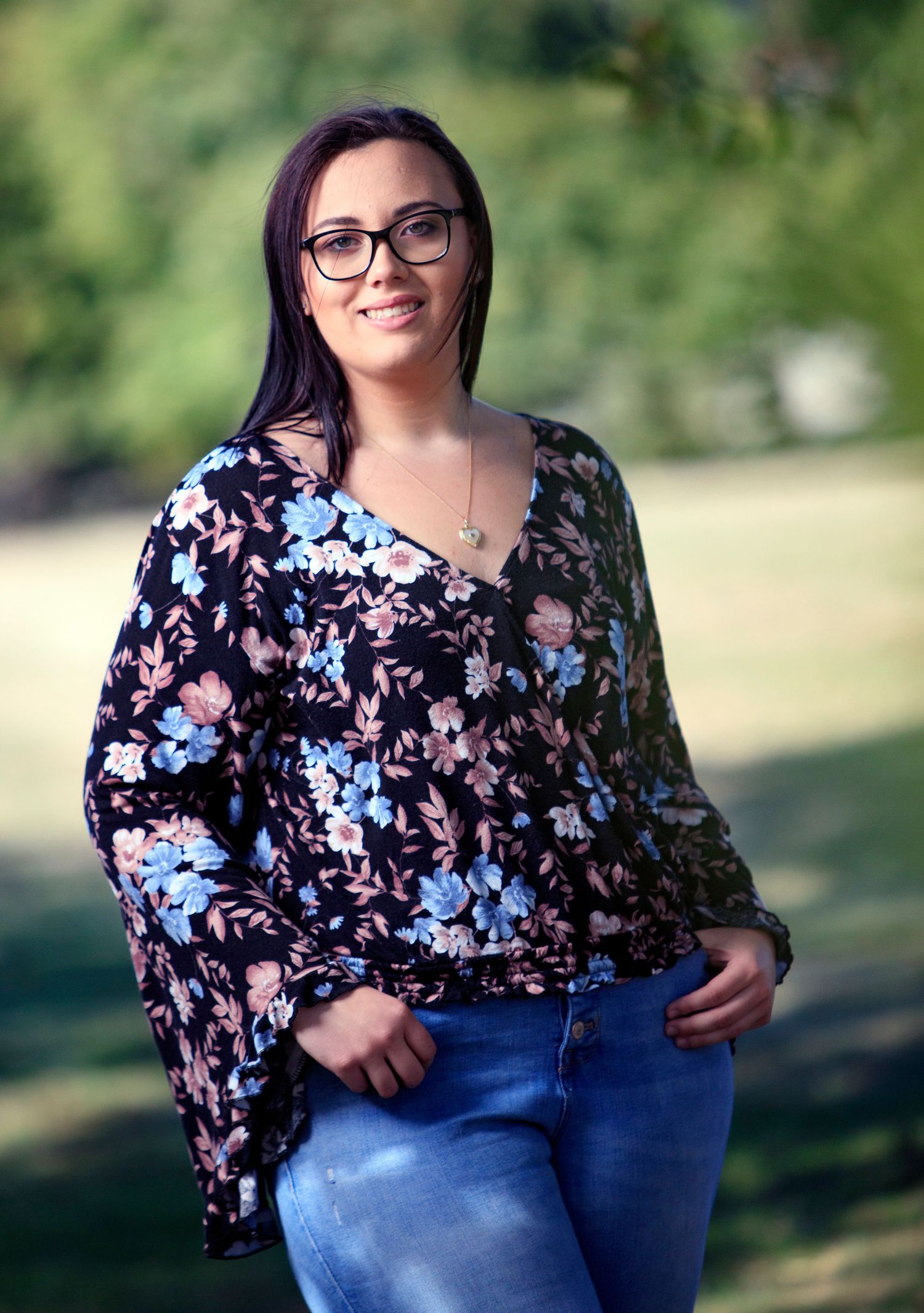 Young woman in floral blouse and jeans smiles while standing in green outdoor park setting.