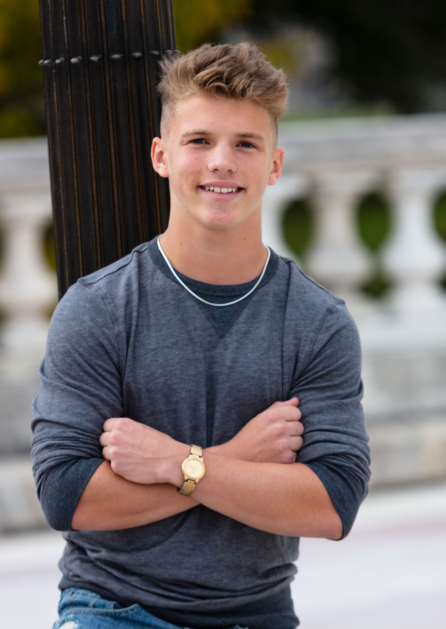 Smiling teen boy in gray sweater with arms crossed leaning on pole in outdoor senior portrait.