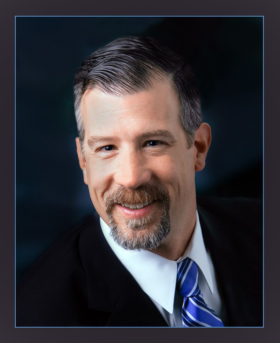 Professional headshot of a smiling middle-aged man with gray hair and goatee wearing a dark suit and striped tie.