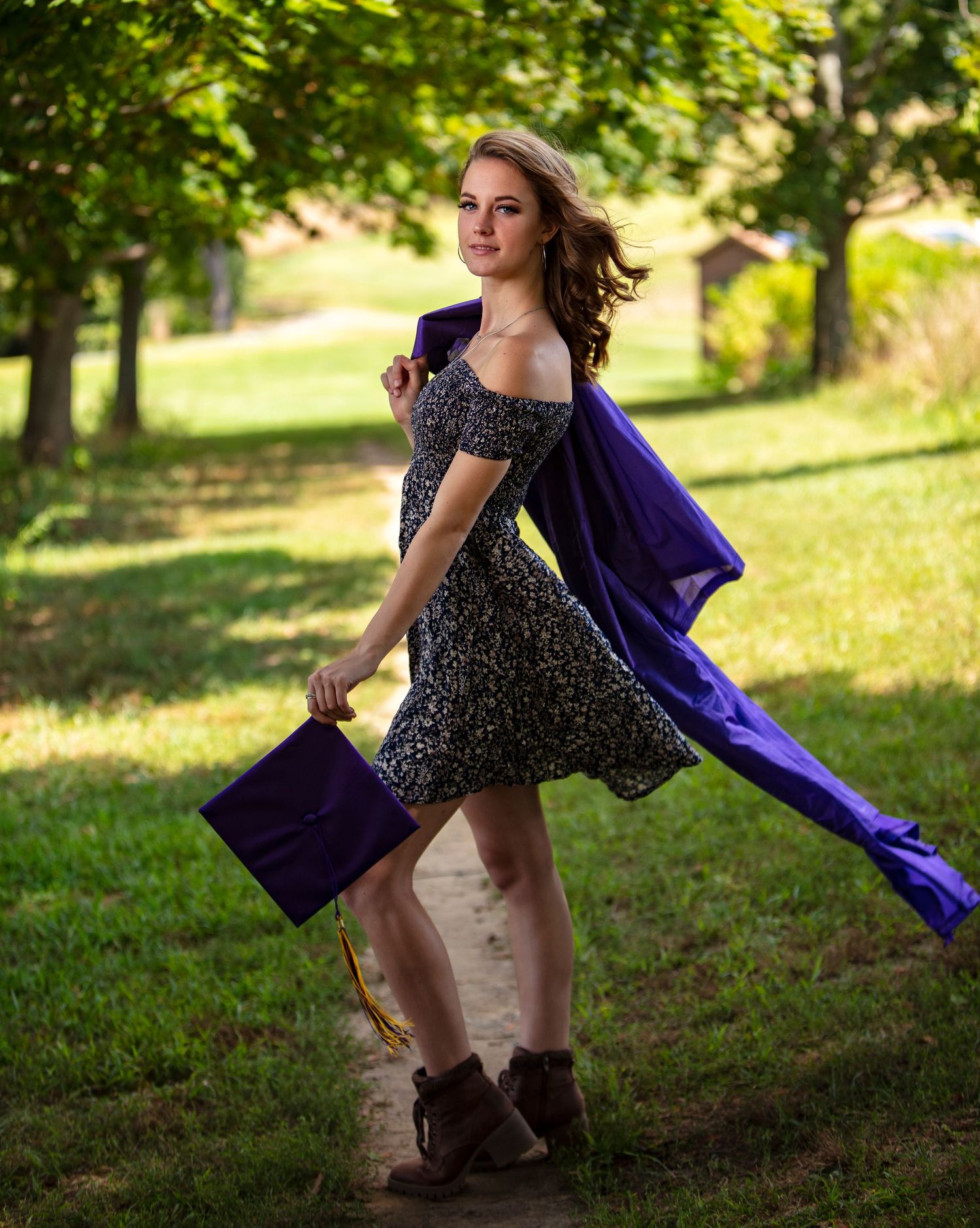 Young woman in floral dress holding purple graduation cap and gown outdoors in green park setting.