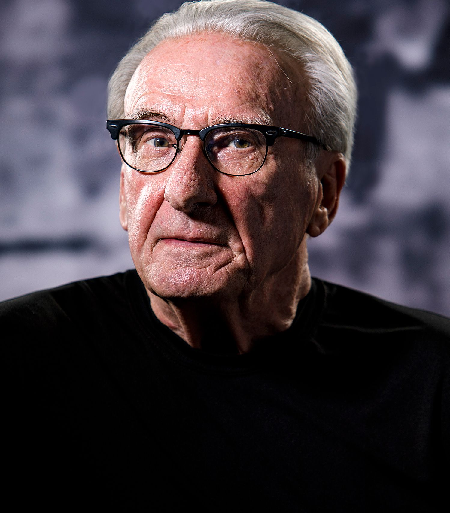 Elderly man with white hair and black-rimmed glasses wearing a black shirt, photographed against a dark background.
