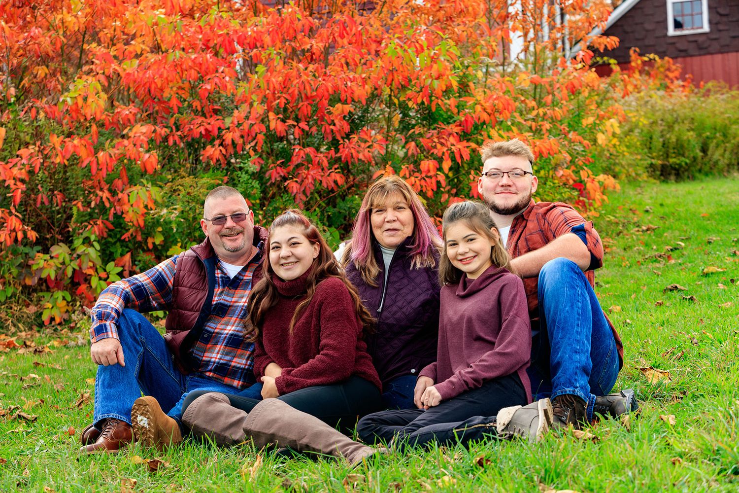 A family of five sits on grass in front of vibrant red autumn foliage.