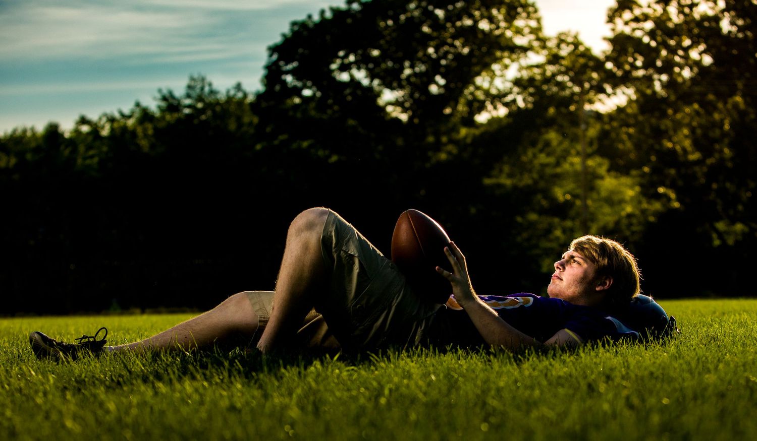 Young man relaxing on green grass holding football in sunny park at golden hour.