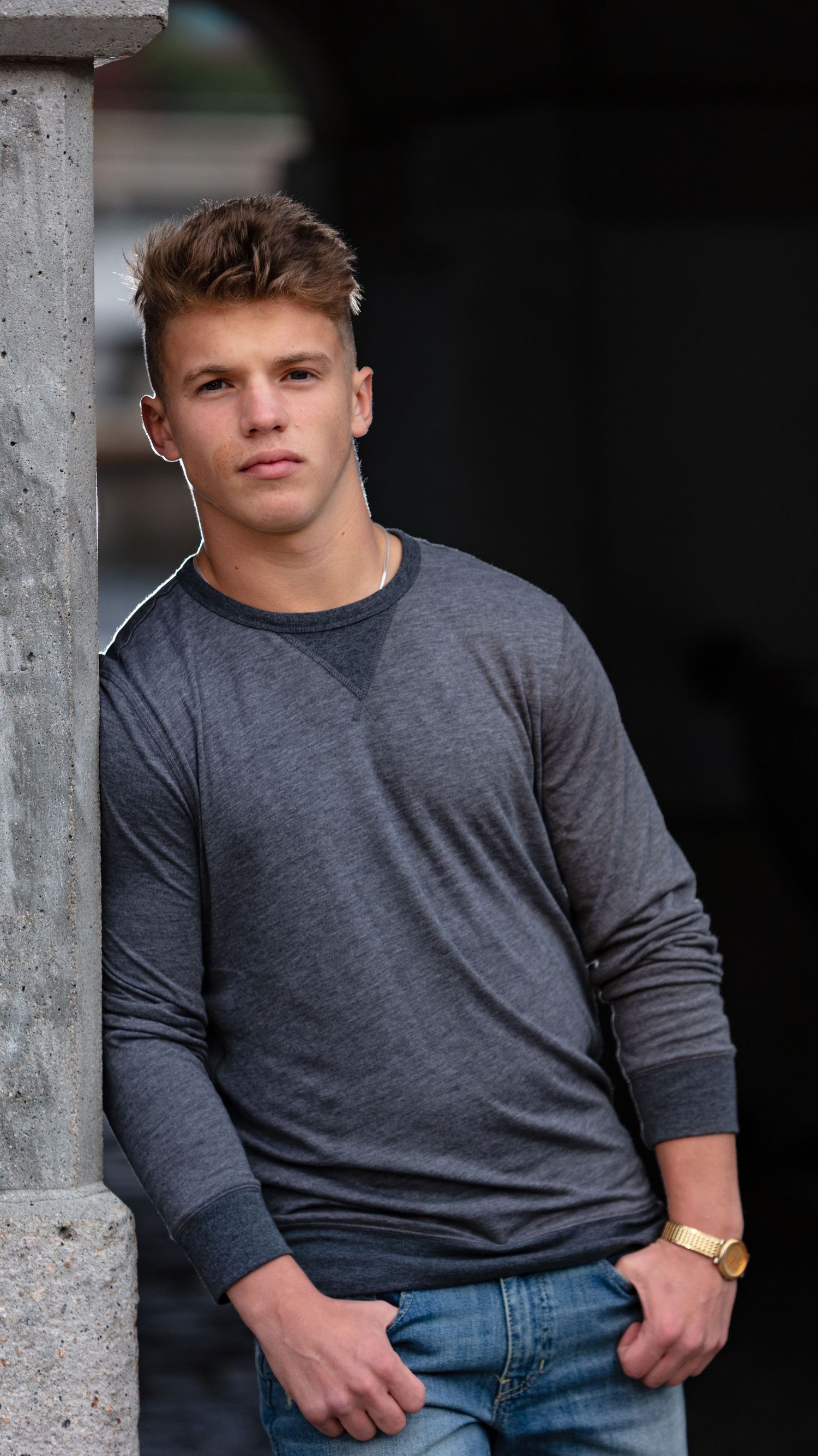 Teen boy in gray sweatshirt leaning against concrete pillar in dramatic senior portrait photo.