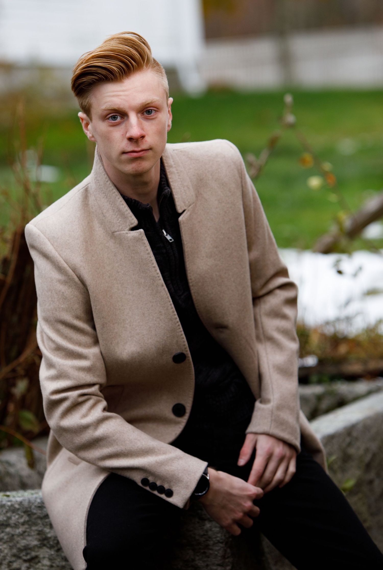 Young man in beige coat and black shirt sitting on stone steps outdoors in autumn setting.