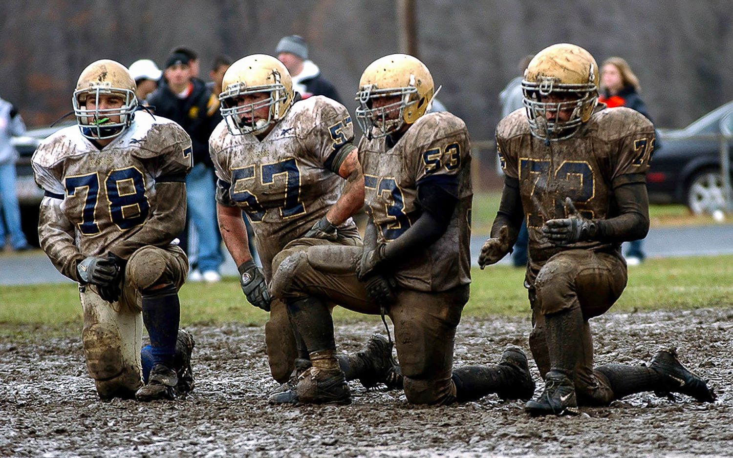 Muddy youth football players in gold uniforms kneel on a wet field during a rainy game day.