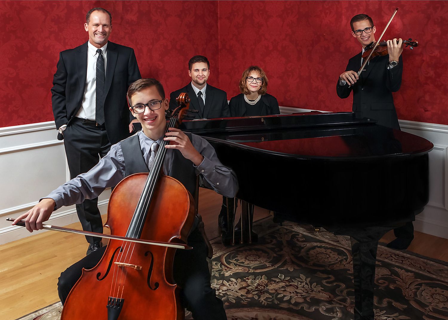 Four classical musicians pose with cello, violin, and grand piano in an elegant red-walled room.