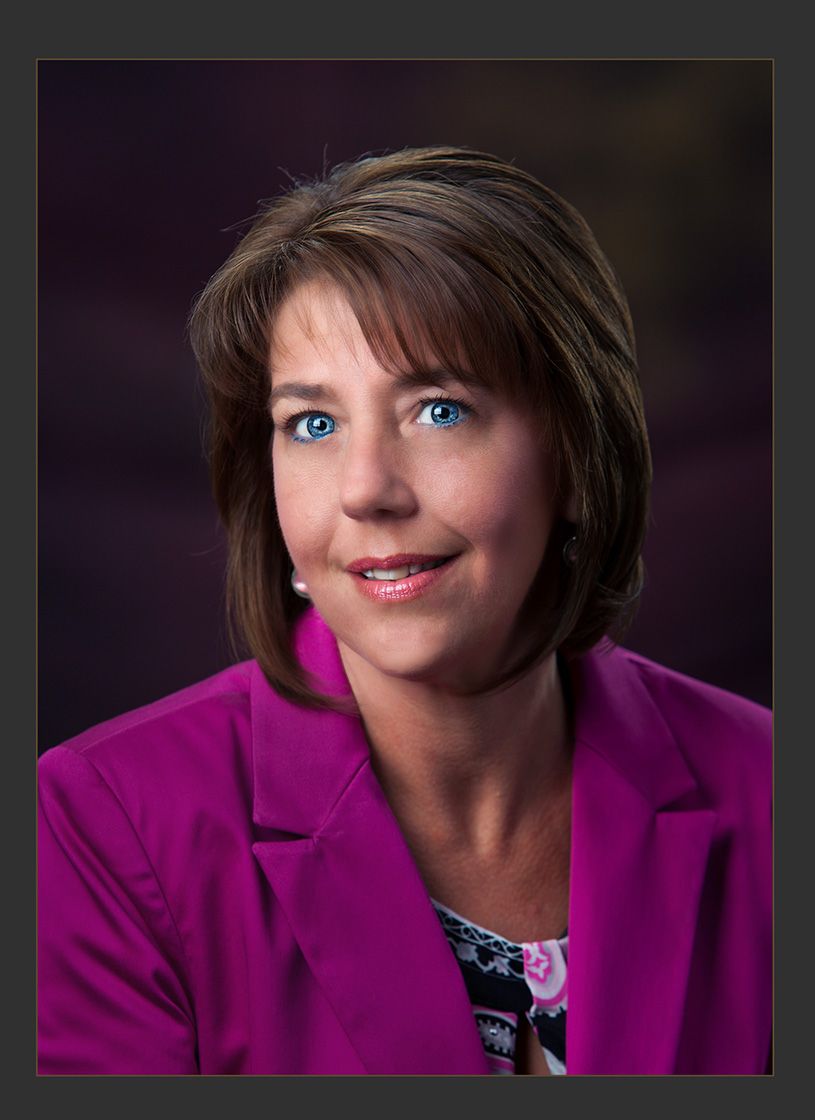 Professional headshot of a smiling woman with brown hair wearing a bright pink blazer against a purple background.