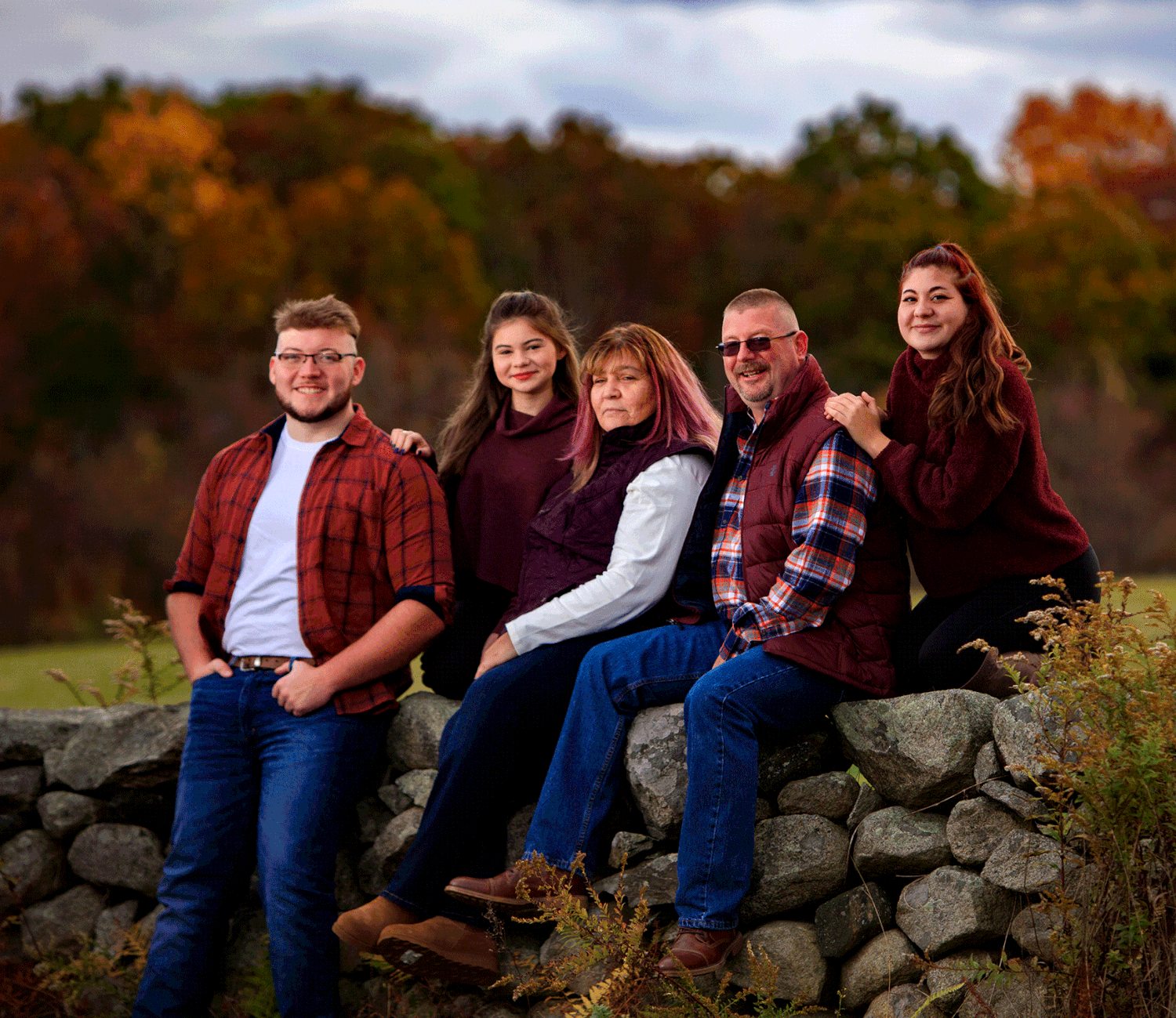 A family of five sits on a stone wall surrounded by colorful autumn foliage.