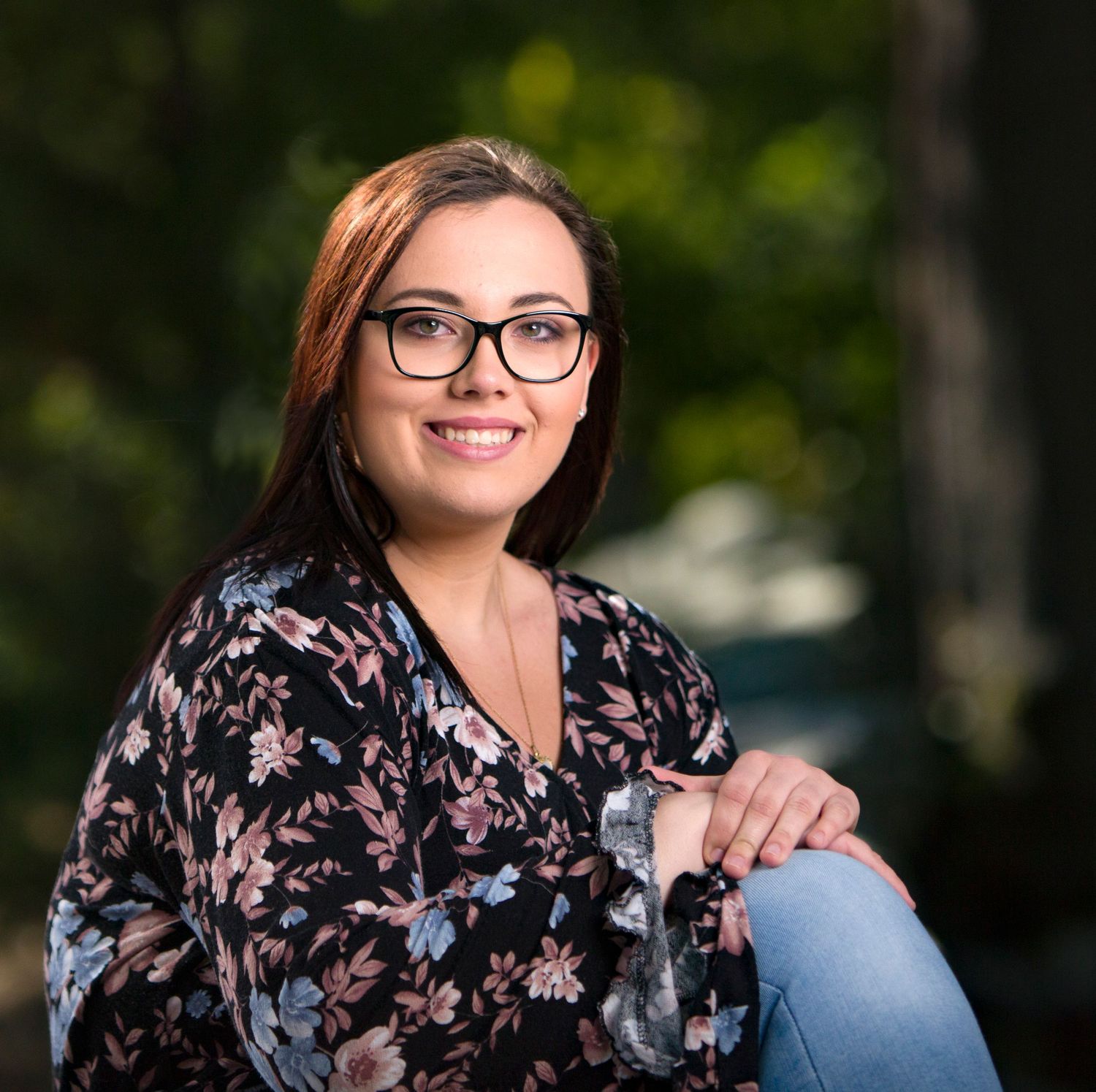 Smiling young woman in floral blouse sits outdoors surrounded by lush green bokeh background.