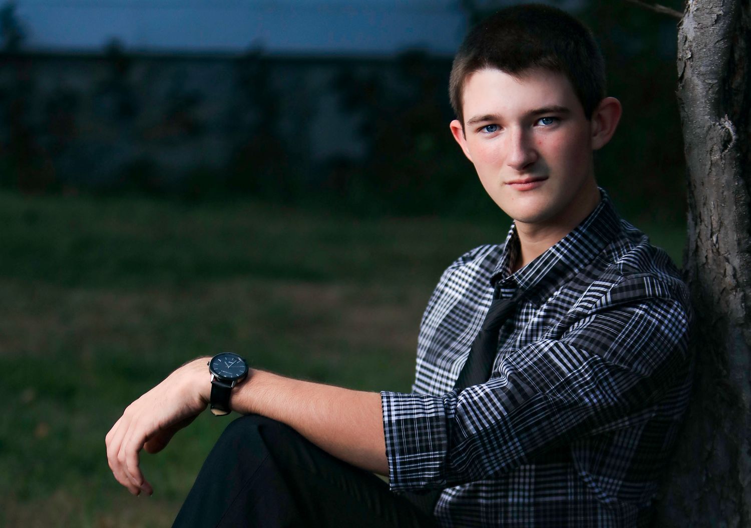 Young man in plaid shirt leaning against tree outdoors, wearing a watch, in a casual portrait photo.