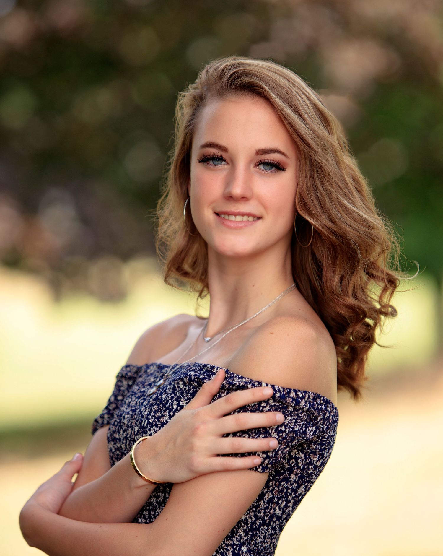 Smiling teenage girl in off-shoulder floral dress with curly hair in sunny park.