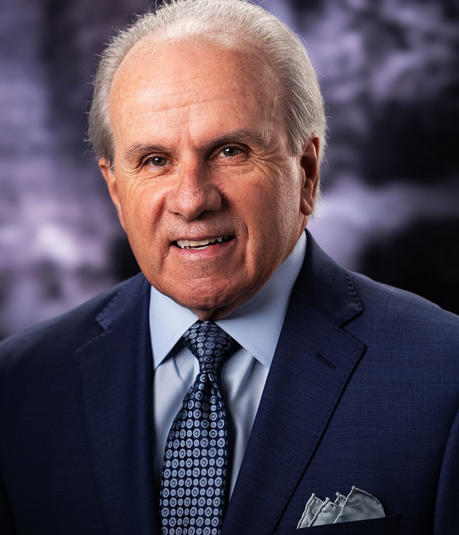 Professional headshot of a senior businessman in a navy suit and patterned tie with gray hair.