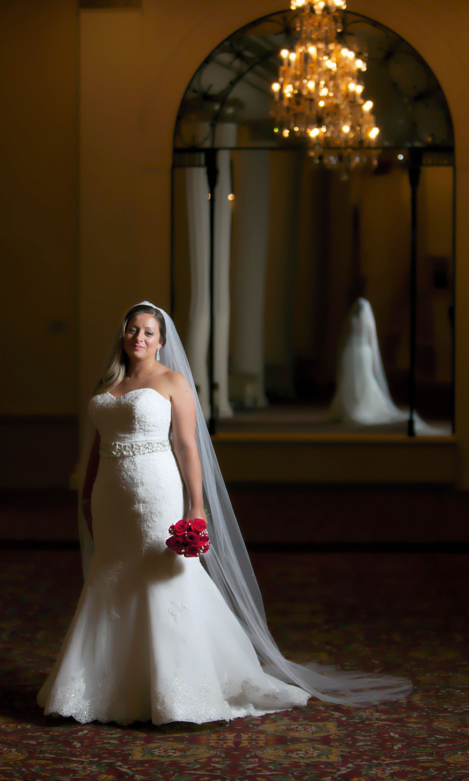 Bride in strapless white mermaid gown with veil holds red bouquet in elegant ballroom with chandelier.