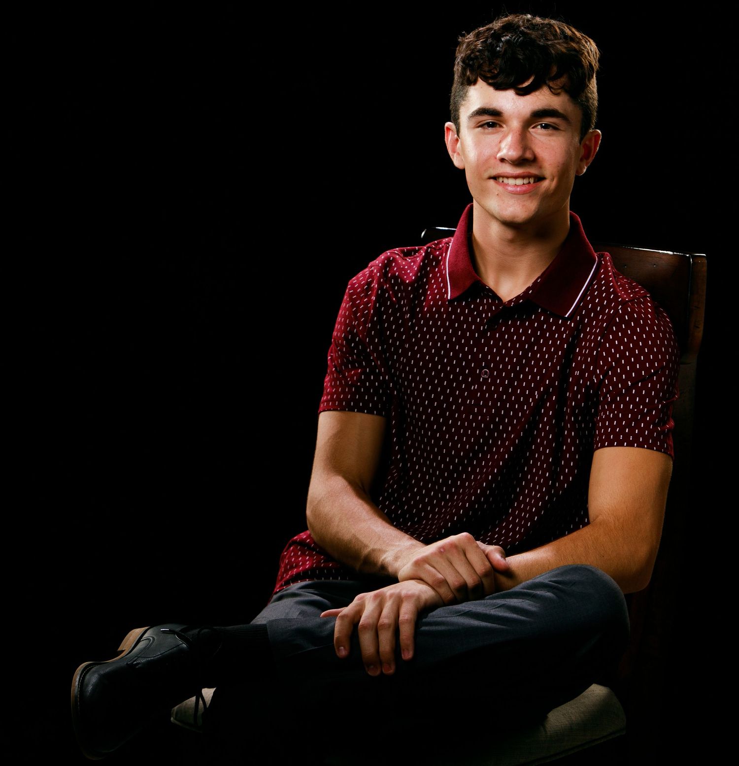 Smiling teenage boy in red polka dot polo shirt seated against dark background.