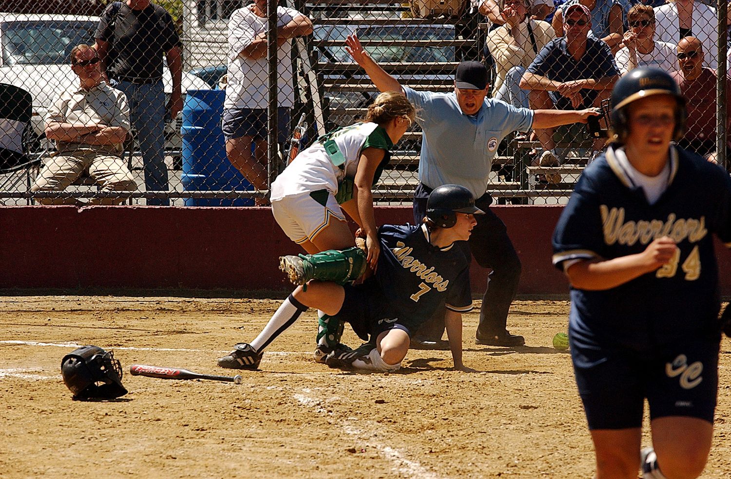 Softball players in action at home plate, with a sliding runner, catcher, and Warriors player on a dirt field.