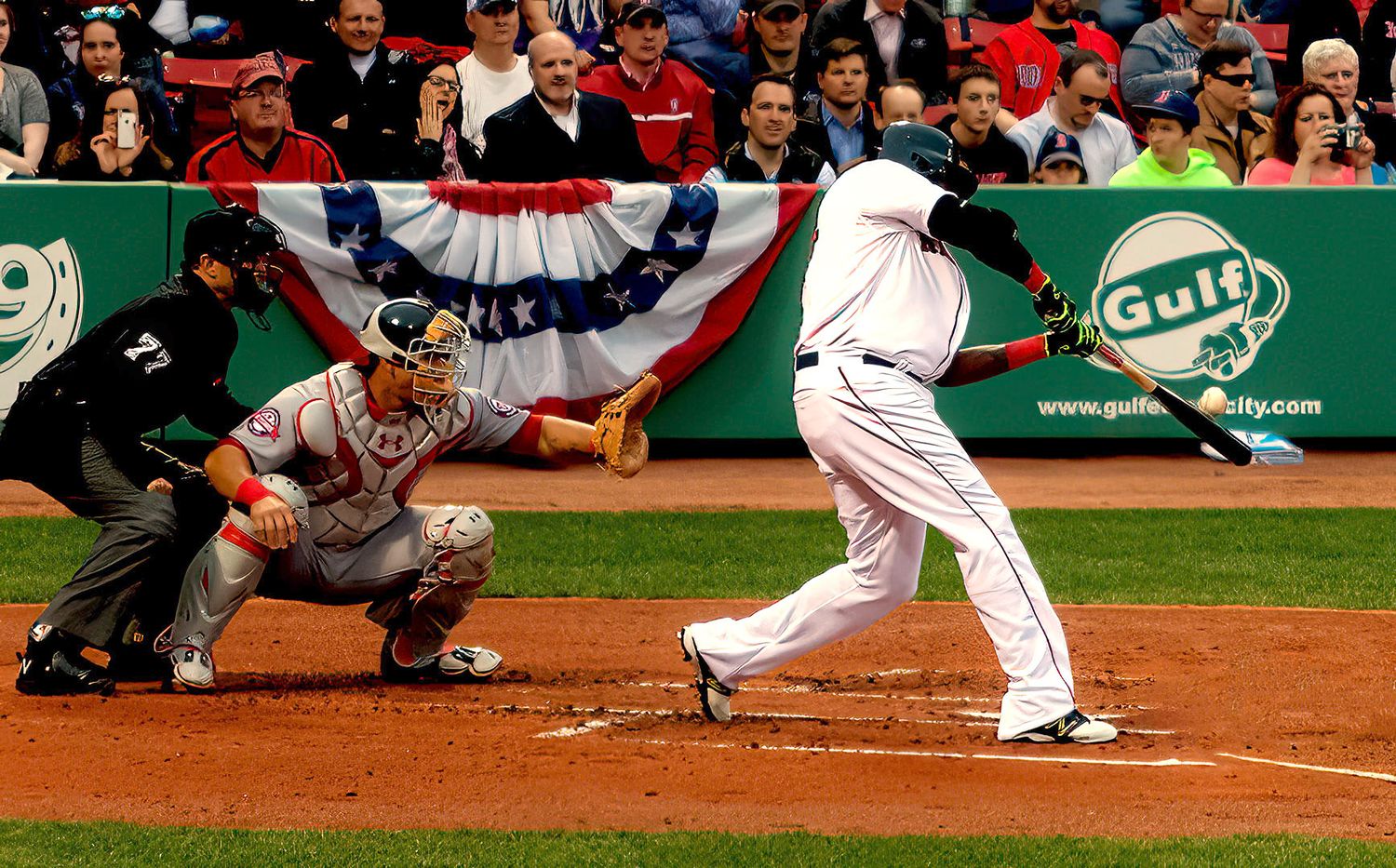 Baseball batter swings at pitch during MLB game with catcher and umpire, patriotic bunting decorates the stadium.