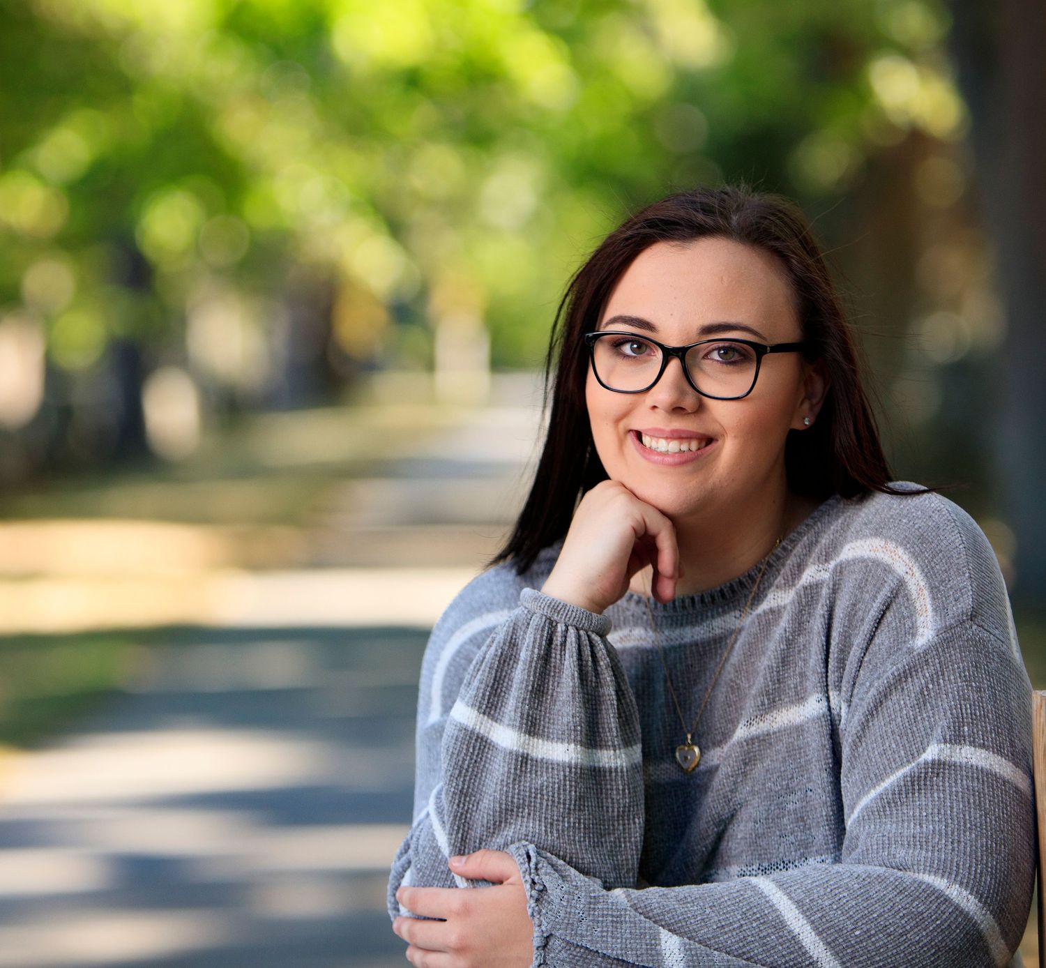 Young woman in gray striped sweater sits smiling outdoors on sunny tree-lined path.