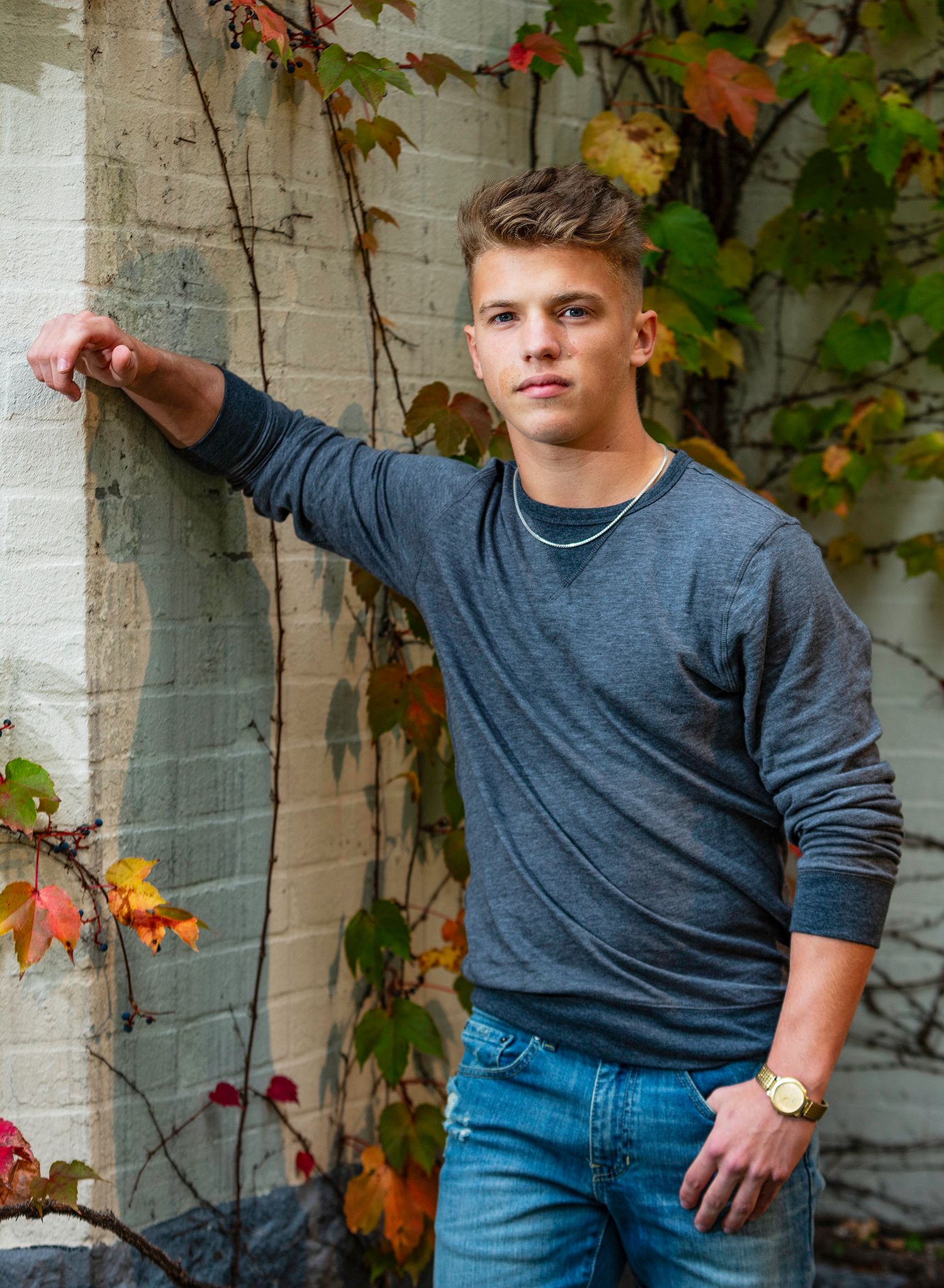 Young man in gray sweater and jeans posing against brick wall with colorful autumn ivy foliage.