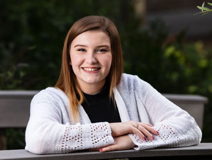 Smiling young woman in white cardigan and black top leaning on a bench outdoors with green foliage background.