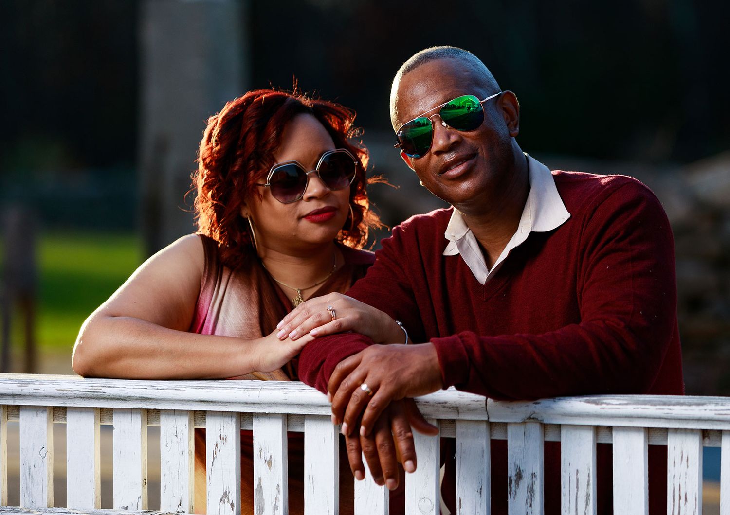 A stylish couple wearing sunglasses leans against a white fence outdoors.