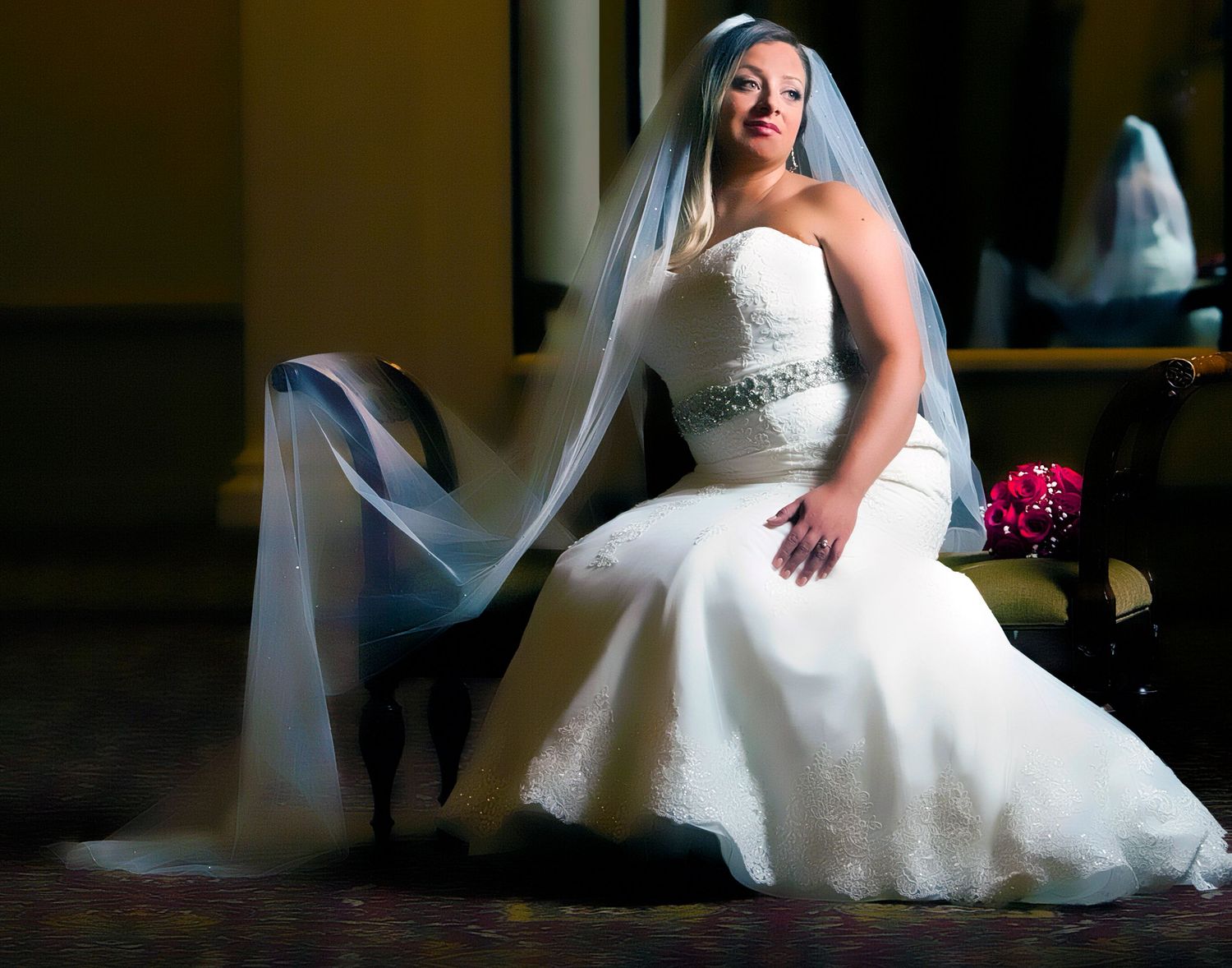 Bride in white strapless wedding gown with veil seated elegantly in chair holding bouquet.