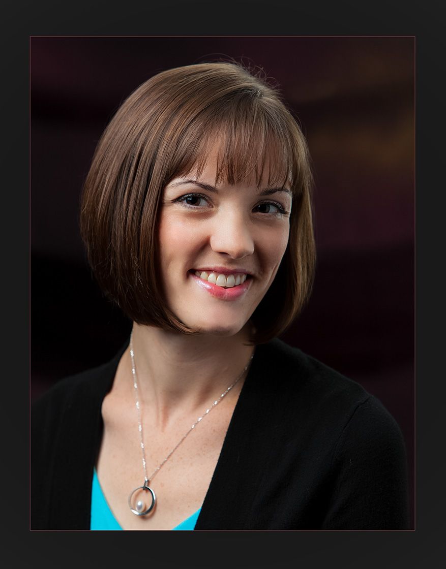 Professional headshot of a smiling woman with a brown bob haircut wearing a black blazer and pendant necklace.