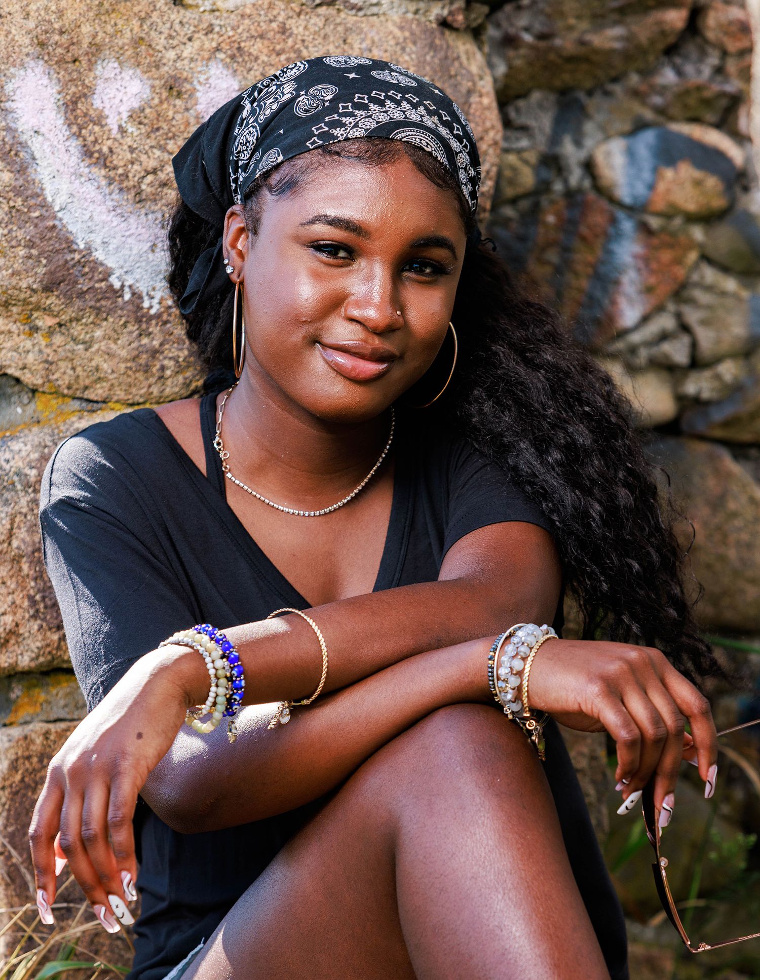Young woman in black outfit with headband and jewelry poses against stone wall outdoors.