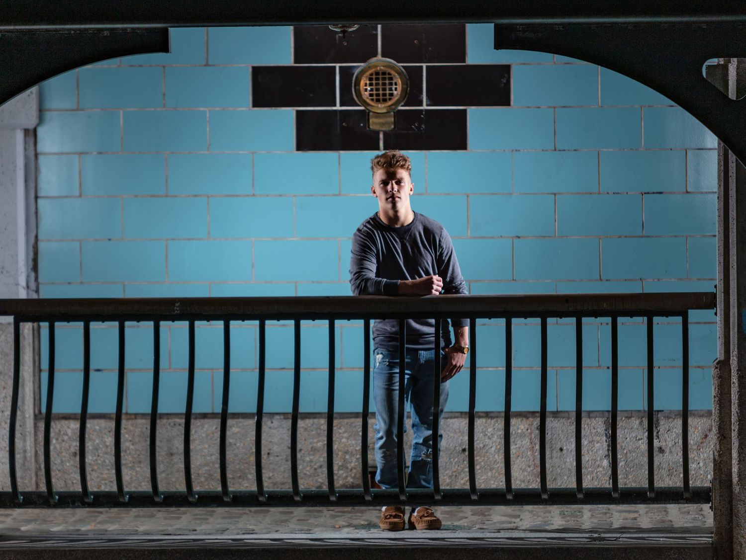 Young man standing at iron railing against blue wall in dramatic nighttime portrait photo.