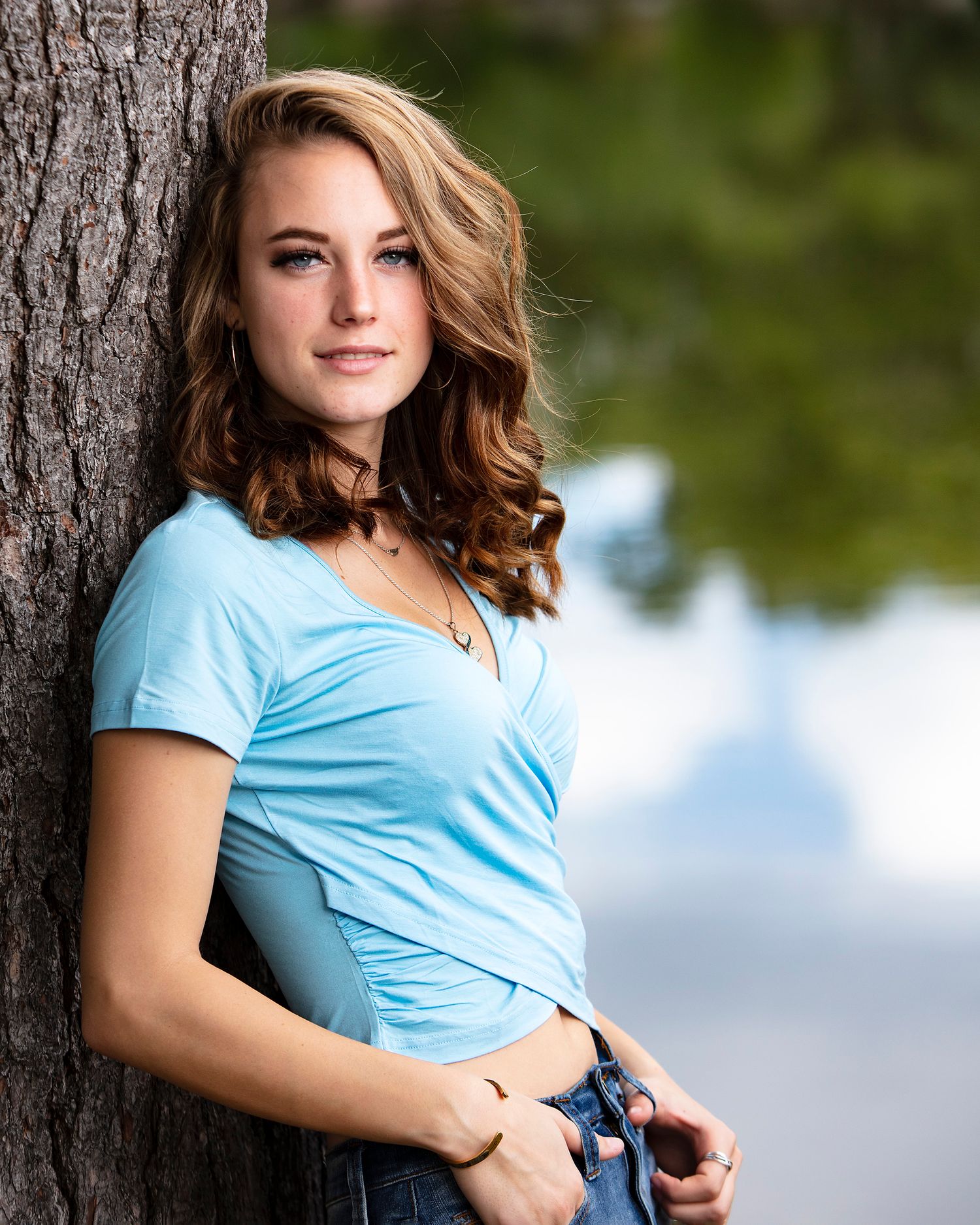 Young woman in light blue top leaning against tree near reflective water, wearing jeans and choker.